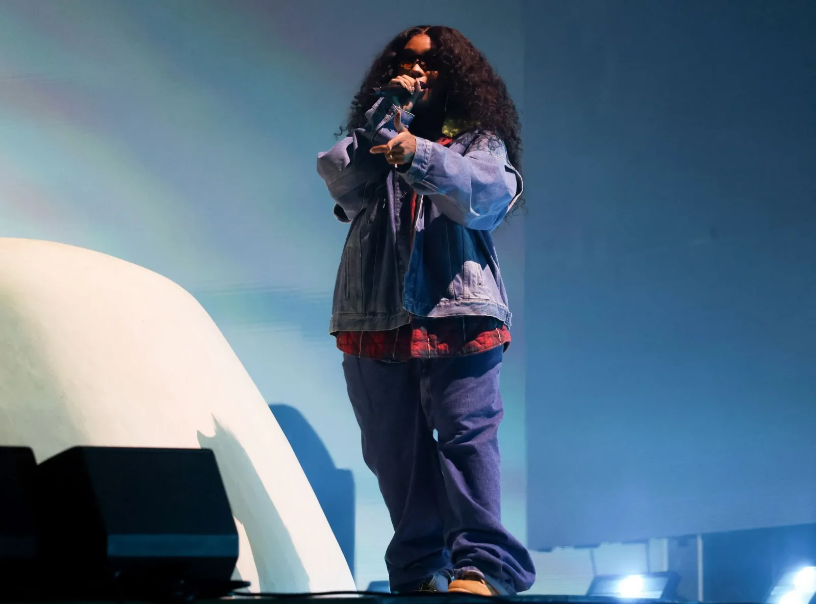 Performer with curly hair and sunglasses raps into a microphone on a blue-lit stage, wearing a denim jacket over a red plaid shirt and baggy jeans.