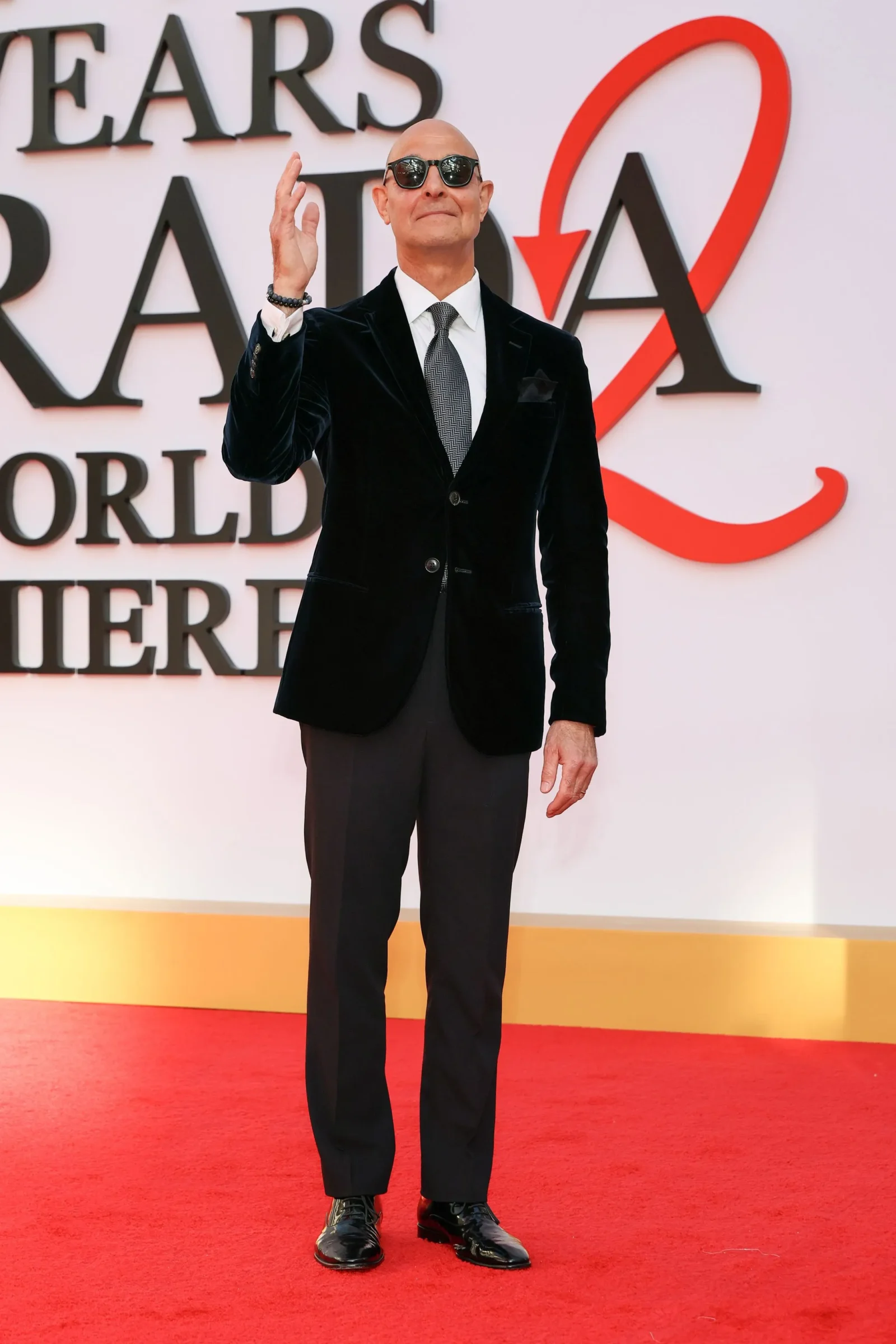 Man in a black velvet suit, white shirt, gray tie, and sunglasses waving on a red carpet at an awards event backdrop.