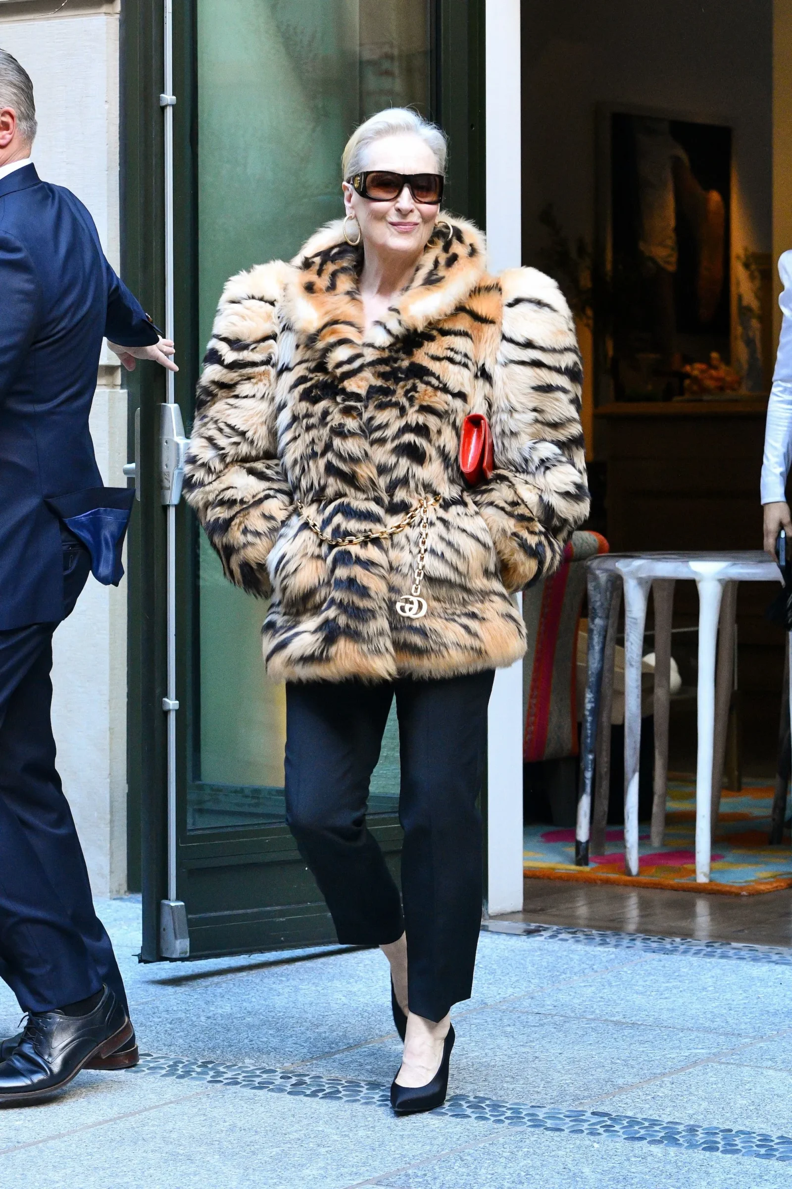 Woman wearing a leopard print fur coat with sunglasses, red clutch, and gold hoops exits a doorway poised and smiling.