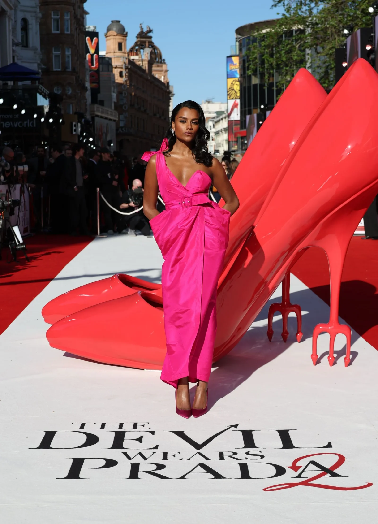 Model in a bright pink dress posing on a red carpet with a large pink sculpture in the background at a fashion event.