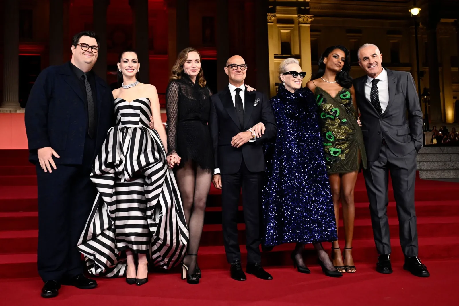 A group of seven well-dressed people posing on red carpet at night, with columned backdrop behind them.