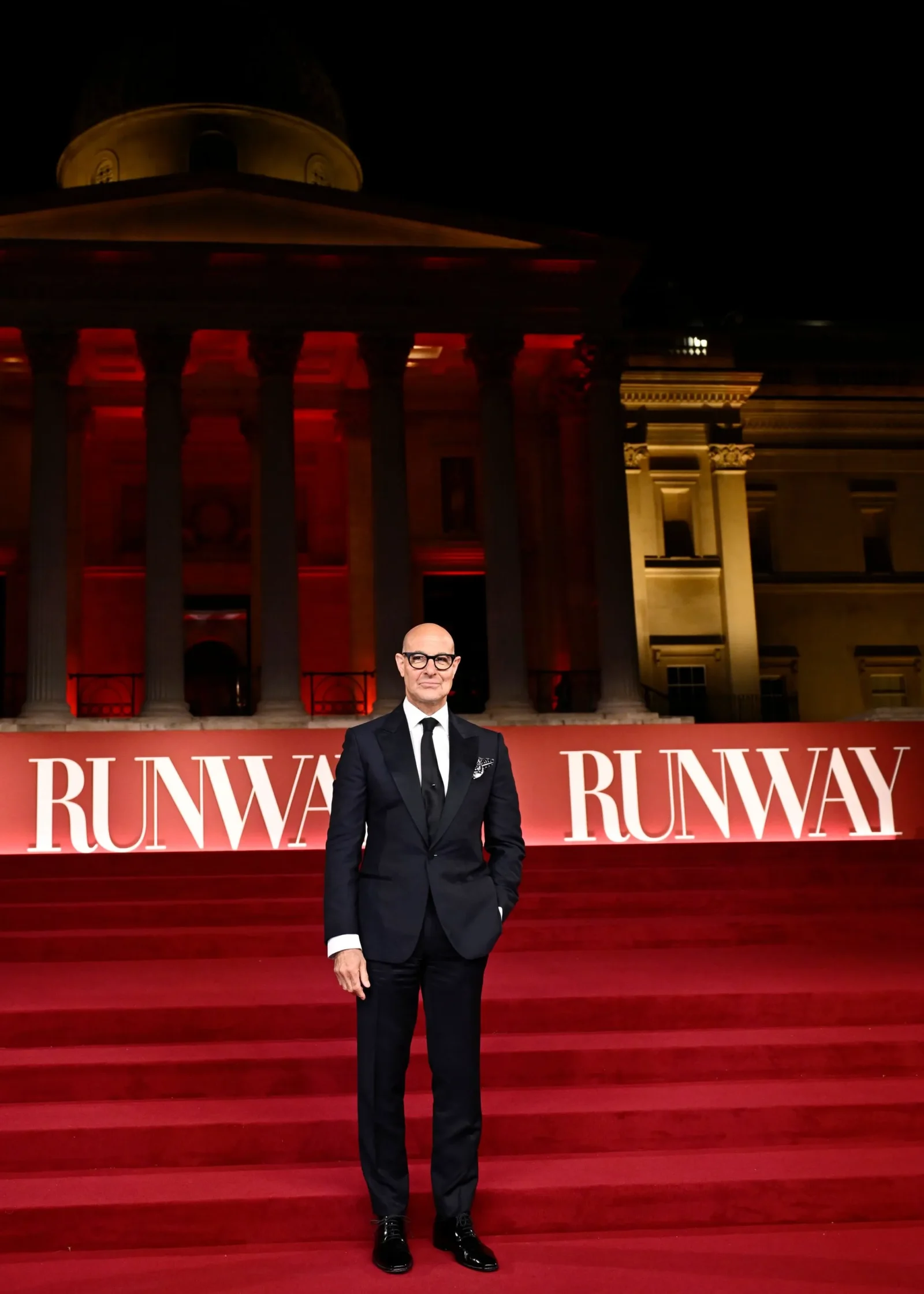 A man in a dark suit and glasses stands on red carpet in front of a grand neoclassical building lit in red and amber, with RUNWAY signs behind him.