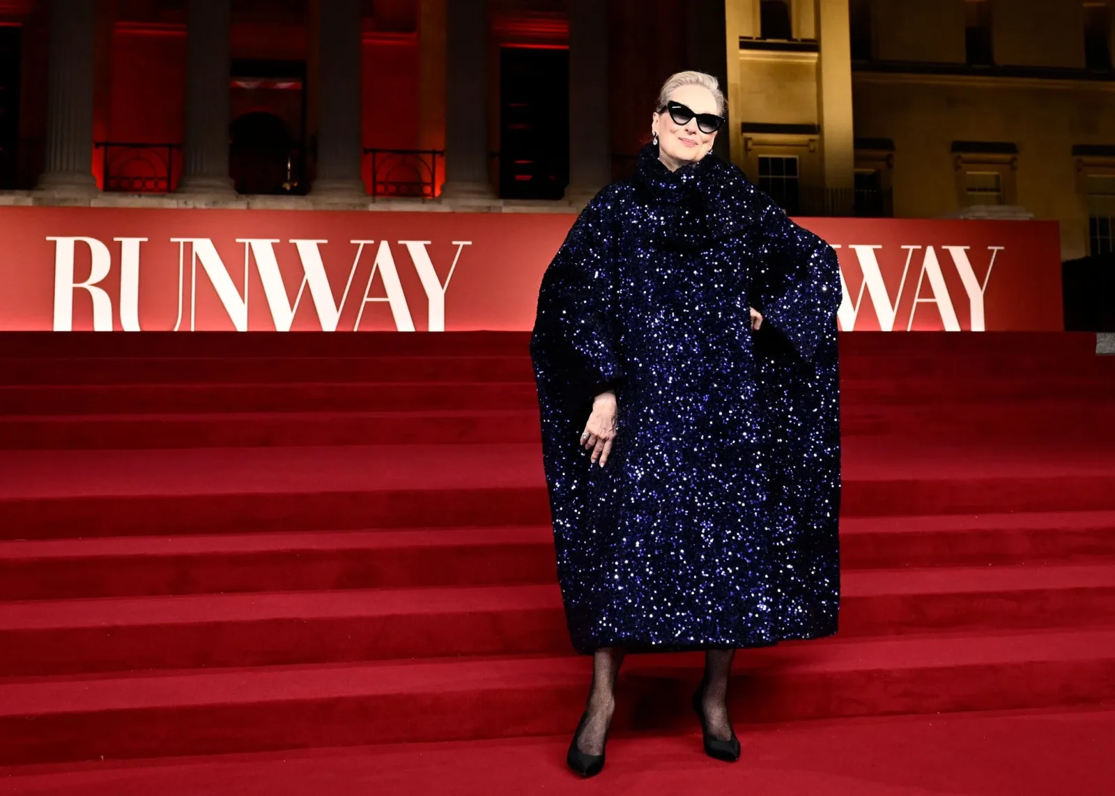 Older woman in a sparkling navy blue sequin gown, sunglasses, posing on a red-carpet runway.