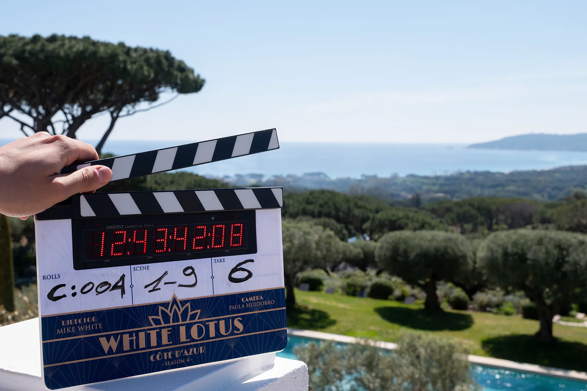 Clapperboard held in the foreground with a coastal landscape behind it, showing the film's title 'WHITE LOTUS' and Côte d'Azur in the design.