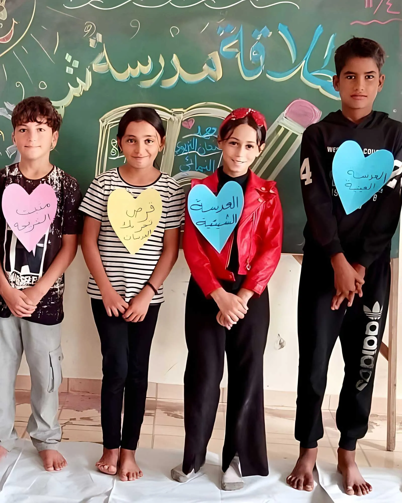 Four students stand in front of a chalkboard, wearing colored paper hearts with Arabic text on their chests.