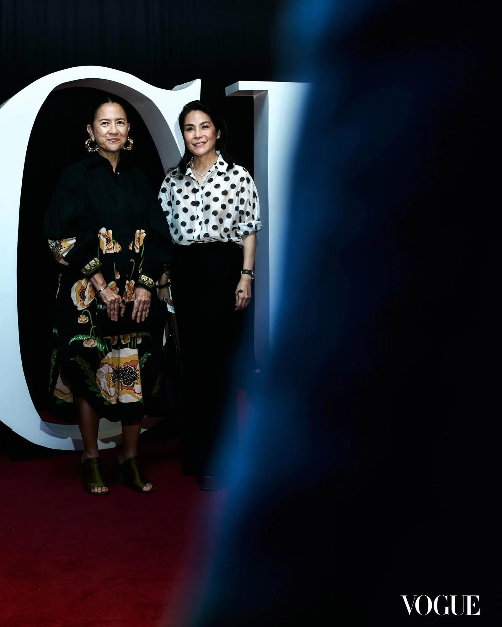 Two women pose in front of a large white sculpture at a Vogue event; one wears a black dress with floral patterns, the other a white polka-dot blouse and black skirt.