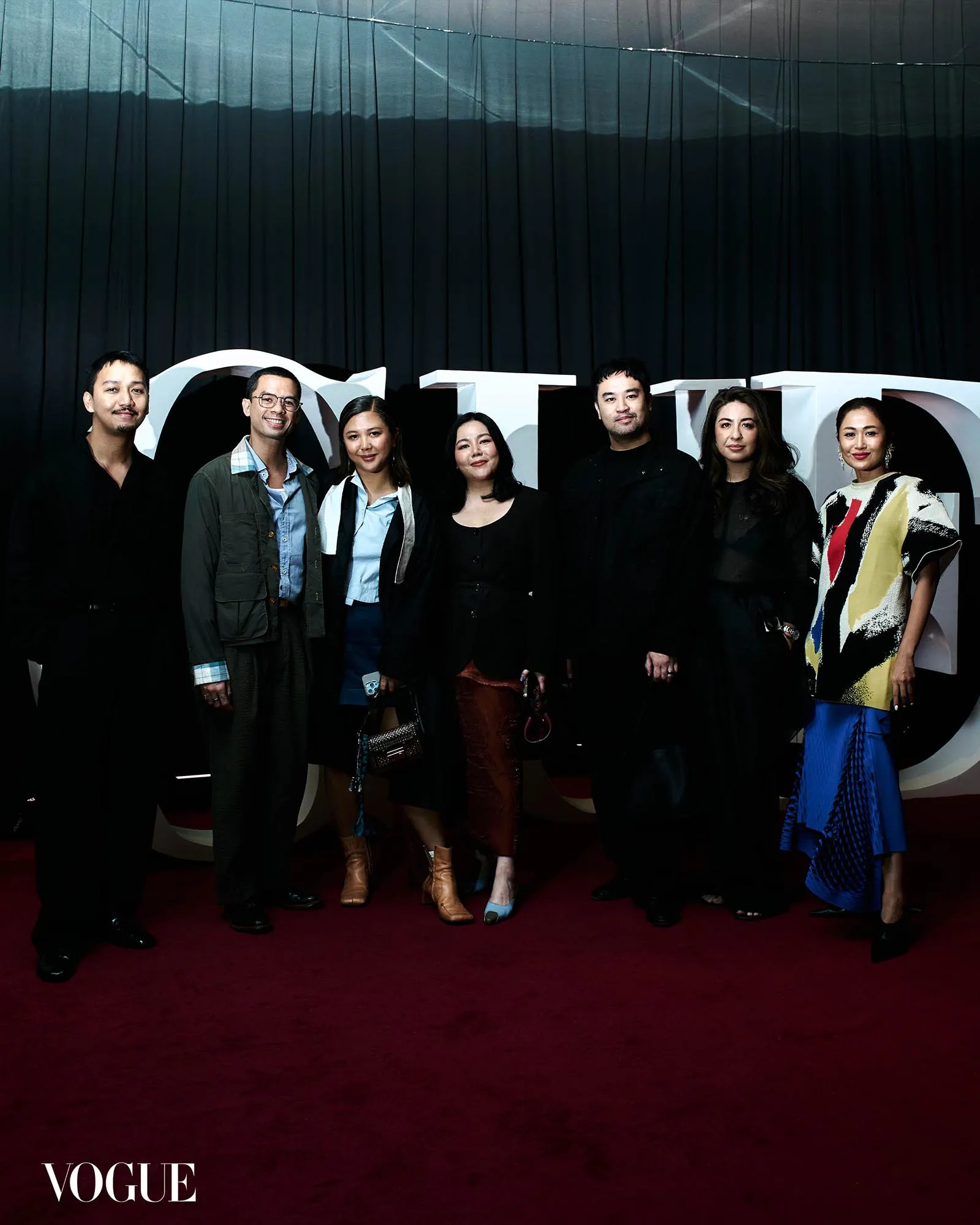 Group of seven people posing on a red carpet at a Vogue event, with large white letters and a dark curtain backdrop behind them.