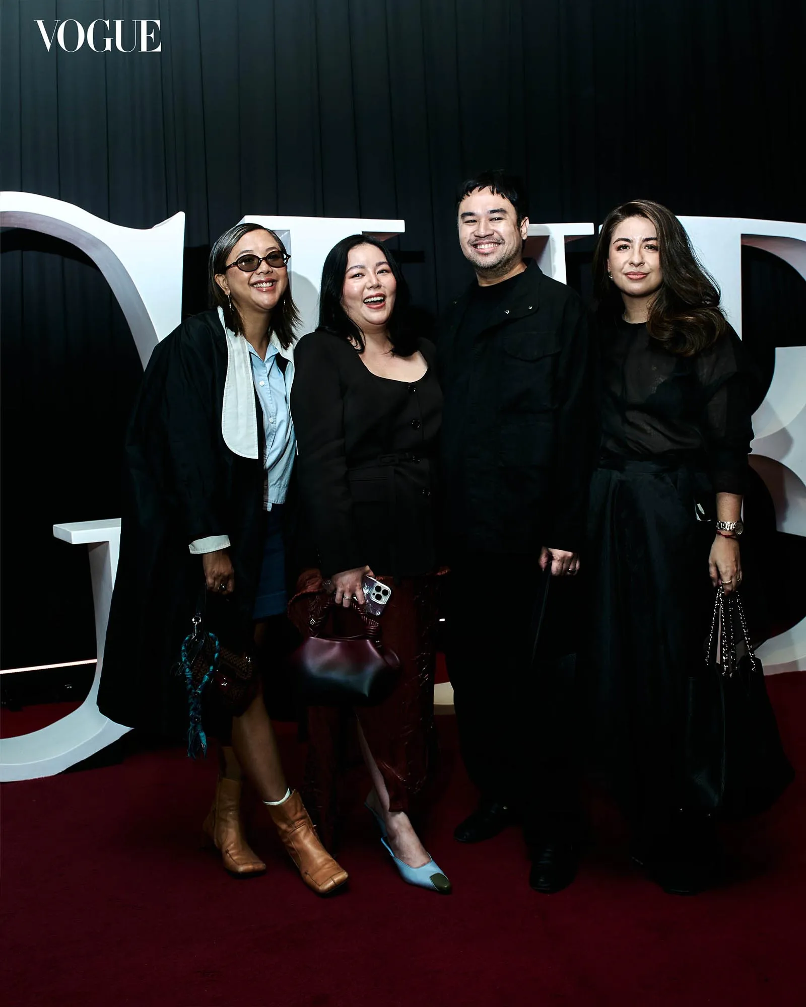 Four people posing and smiling on a red carpet against a black backdrop with Vogue logo behind them.