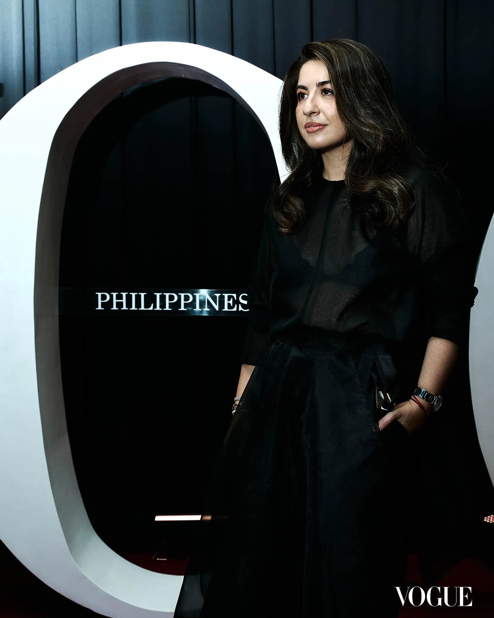 A woman in a black dress posing beside a large white circular sculpture, with the Vogue logo visible in the lower right corner.