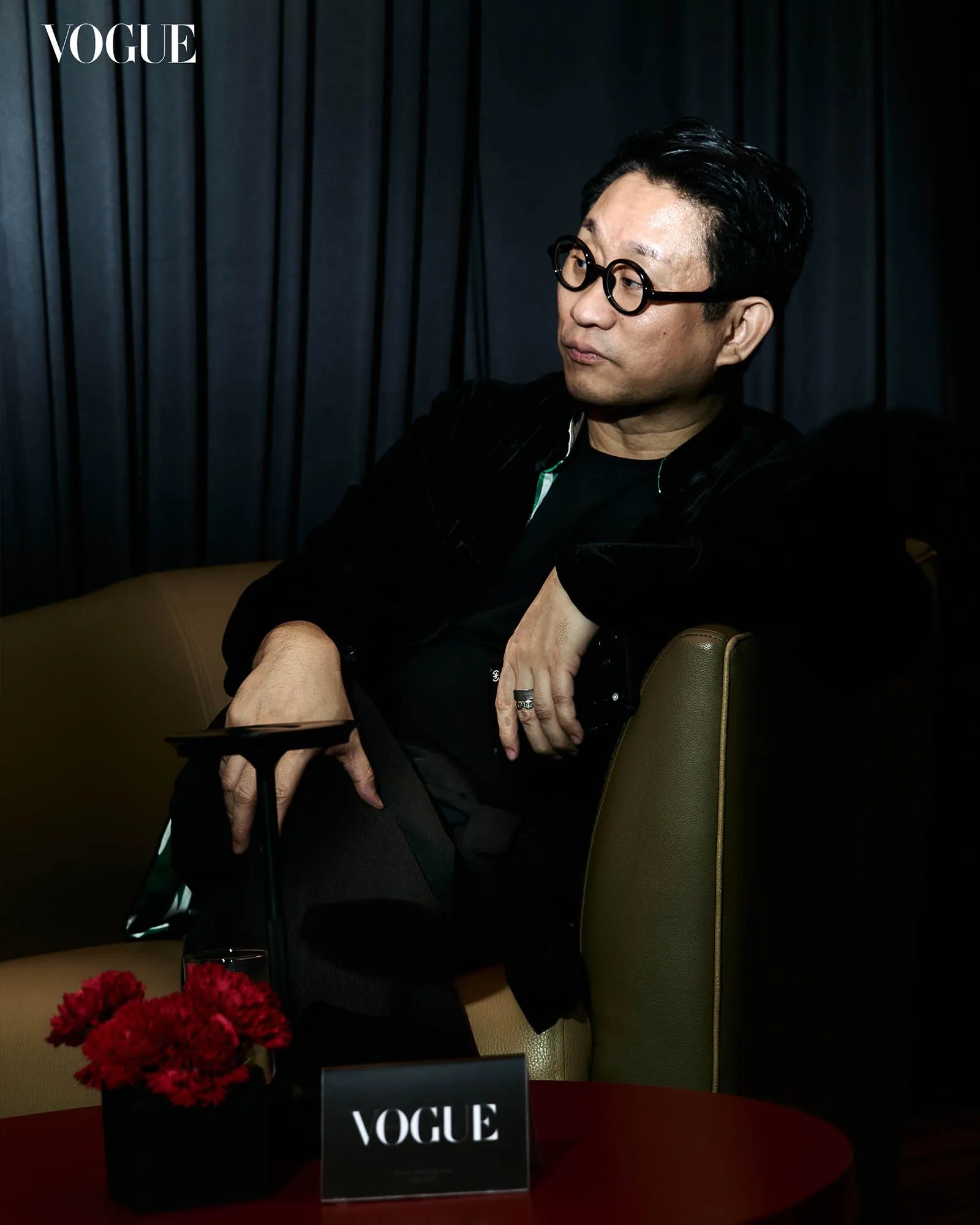 Man with round black glasses sits on a brown chair in front of dark curtains; Vogue logo in the upper left and red flowers on a table in front.