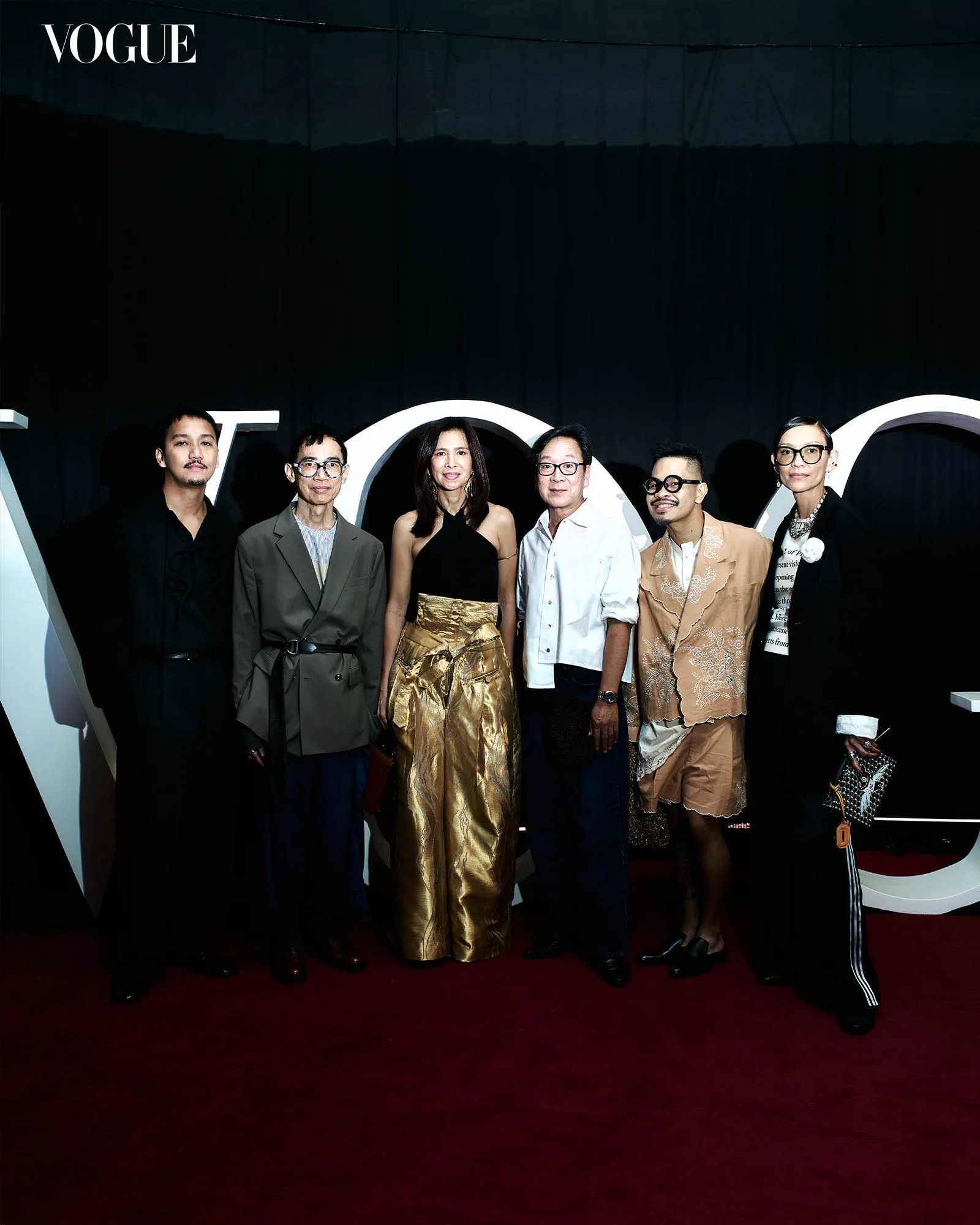 Seven fashionably dressed individuals posing together on a red carpet at a Vogue event, with a dark backdrop behind them.