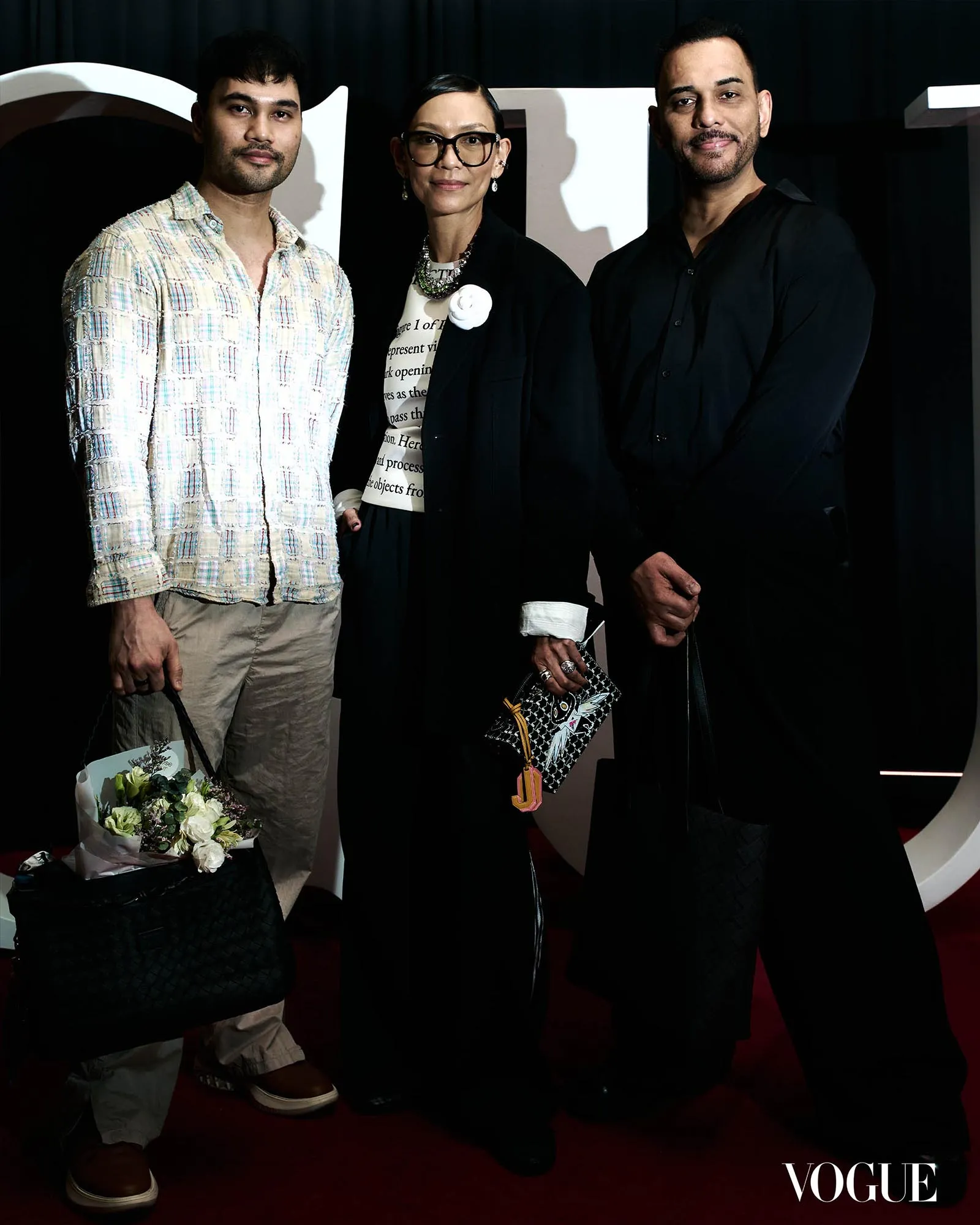 Three fashionably dressed people pose together on a Vogue event backdrop; left holds a bouquet and bag, center wears glasses and a white flower accessory, all smiling.