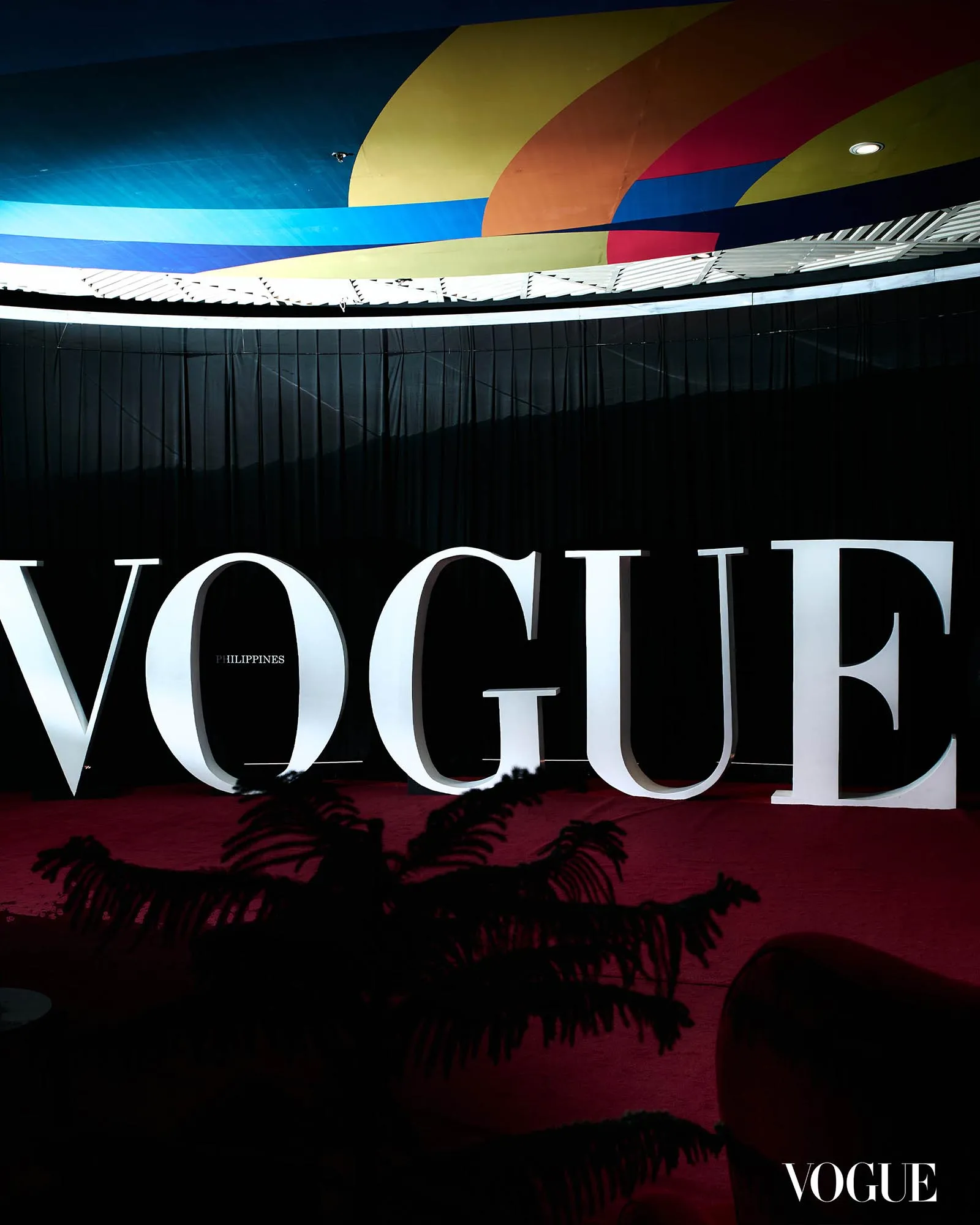 Large white 'VOGUE' letters on a dark backdrop with a colorful curved ceiling design above.