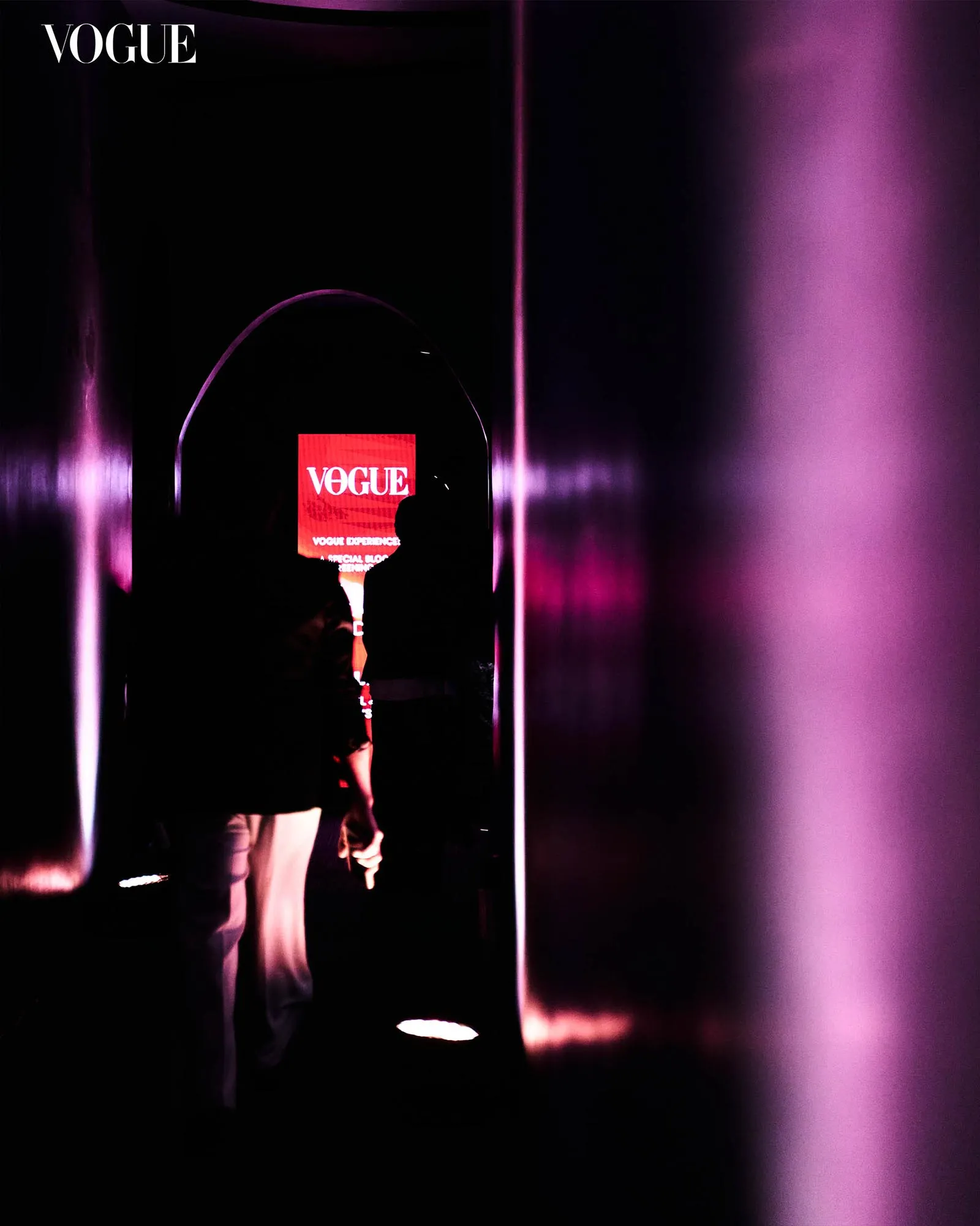 Dark, neon-lit hallway with pink-purple lighting and a Vogue display screen showing the logo; silhouetted attendees in the foreground
