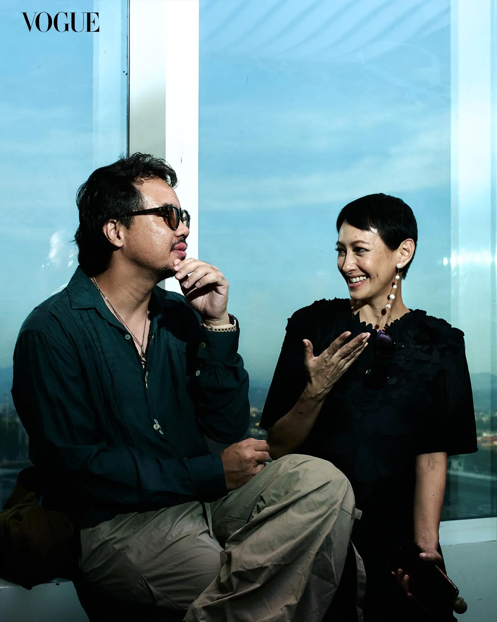 Man and woman engaged in a conversation, seated against a blue glass backdrop with a Vogue logo in the top left corner.