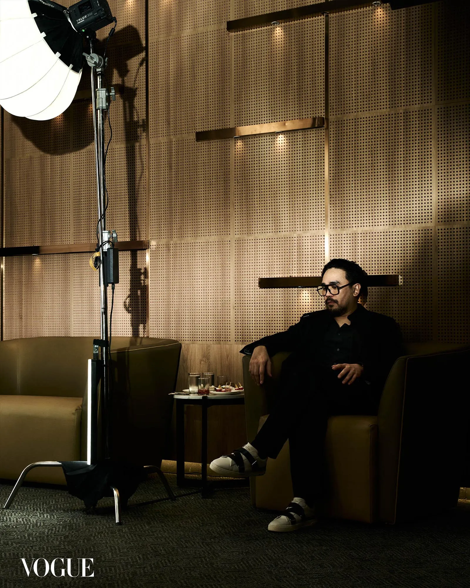 Man in black seated on a sofa in a studio lounge, with photography equipment on the left and a Vogue magazine logo at the bottom corner.