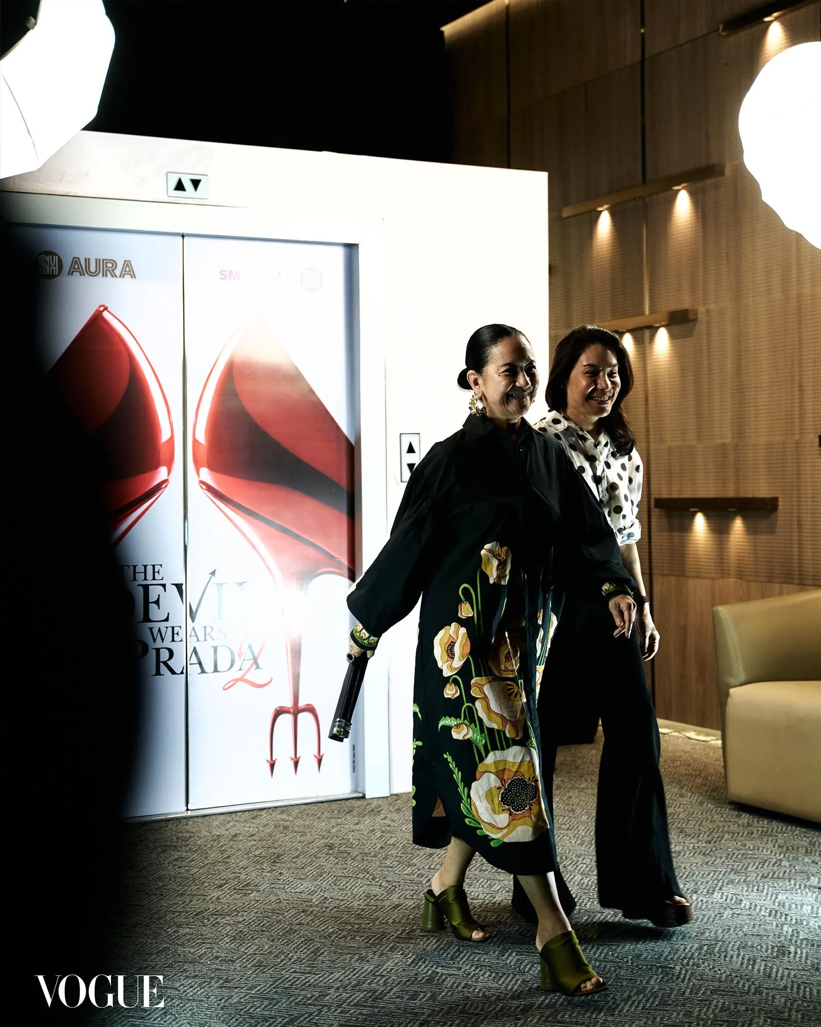 Two smiling women walk past a fashion display with a large Vogue logo in the corner and bright lighting.
