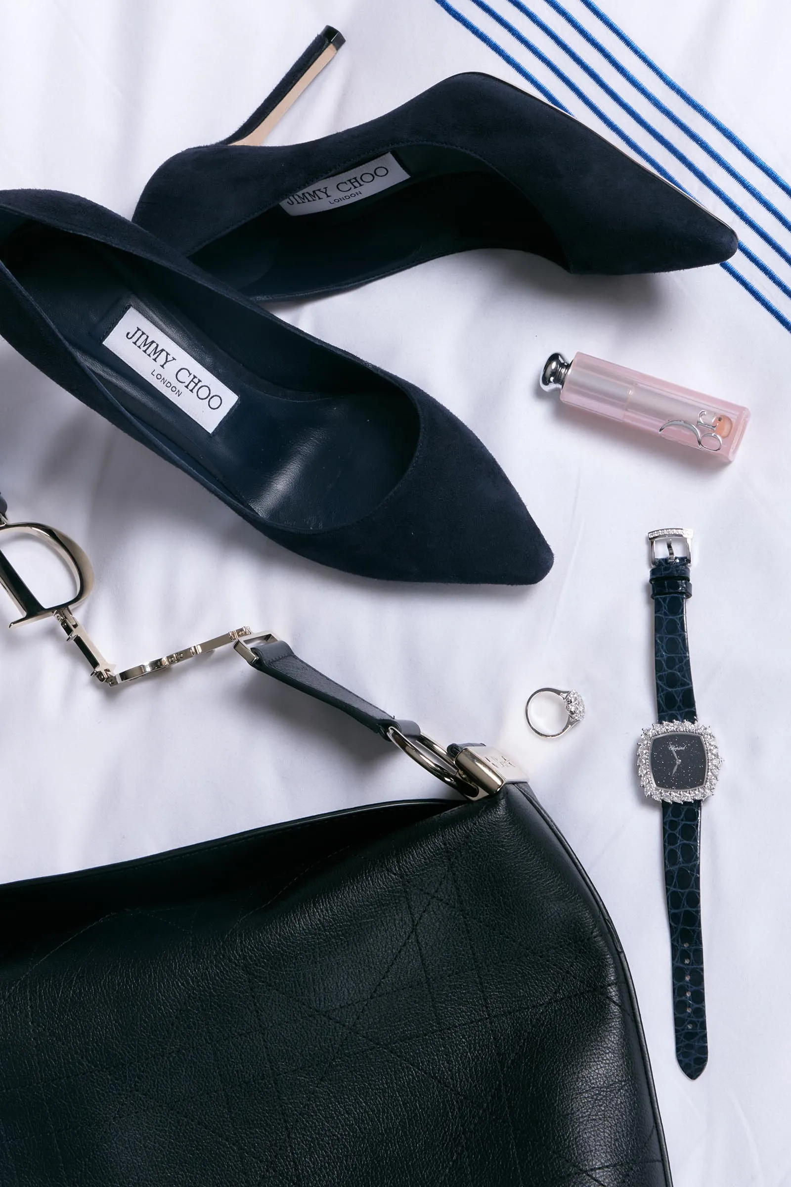 Navy suede Jimmy Choo high heels with a black bag and assorted jewelry on a white surface.