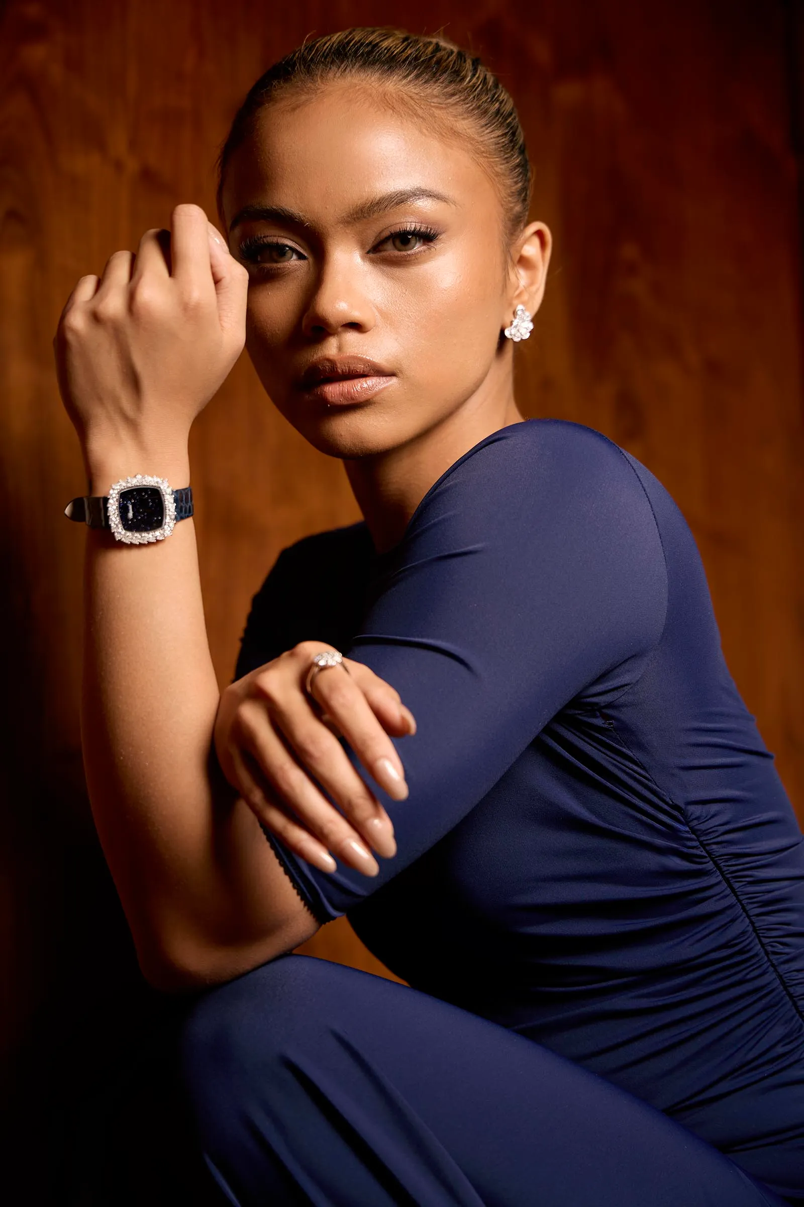 Portrait of a woman in a navy blue dress with diamond earrings and a matching bracelet, posed with her hand near her face against a wooden background.