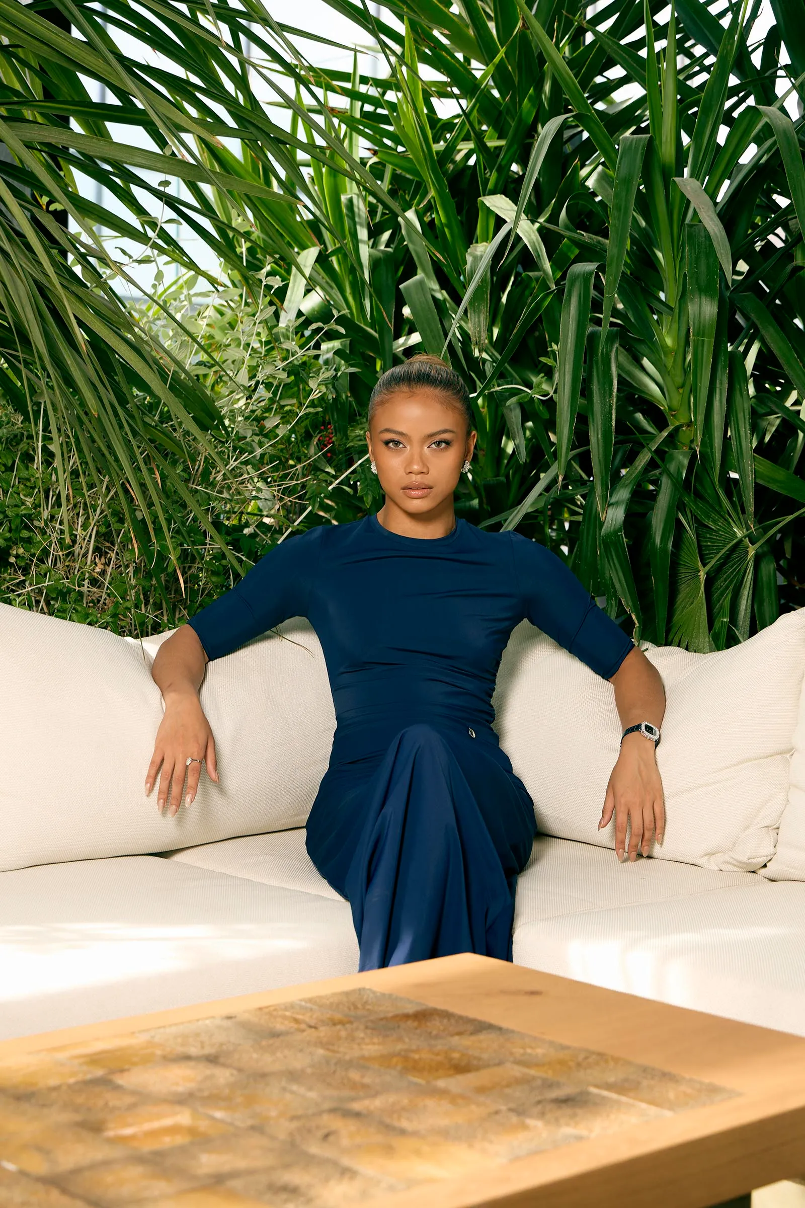 Woman in a navy dress sits on a light sofa with tall green tropical plants in the background.