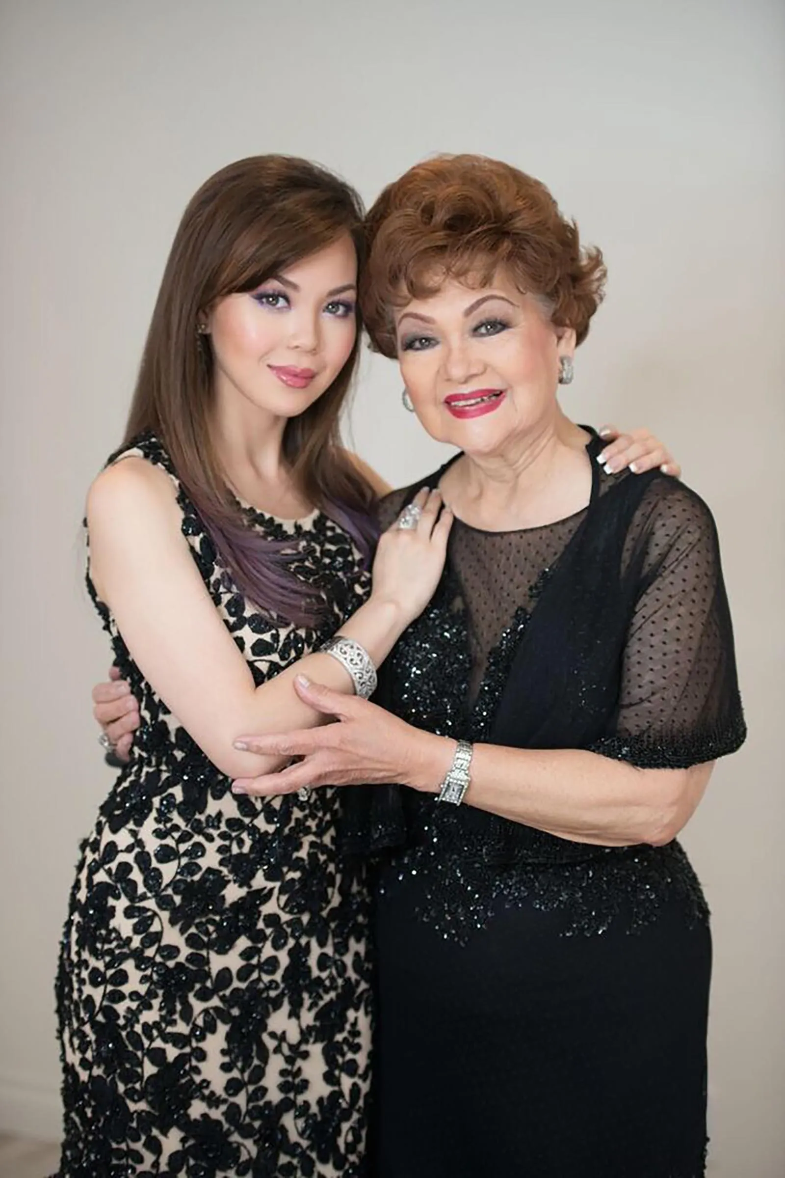 Two women in elegant black dresses hug and smile at the camera, showing warmth and connection.