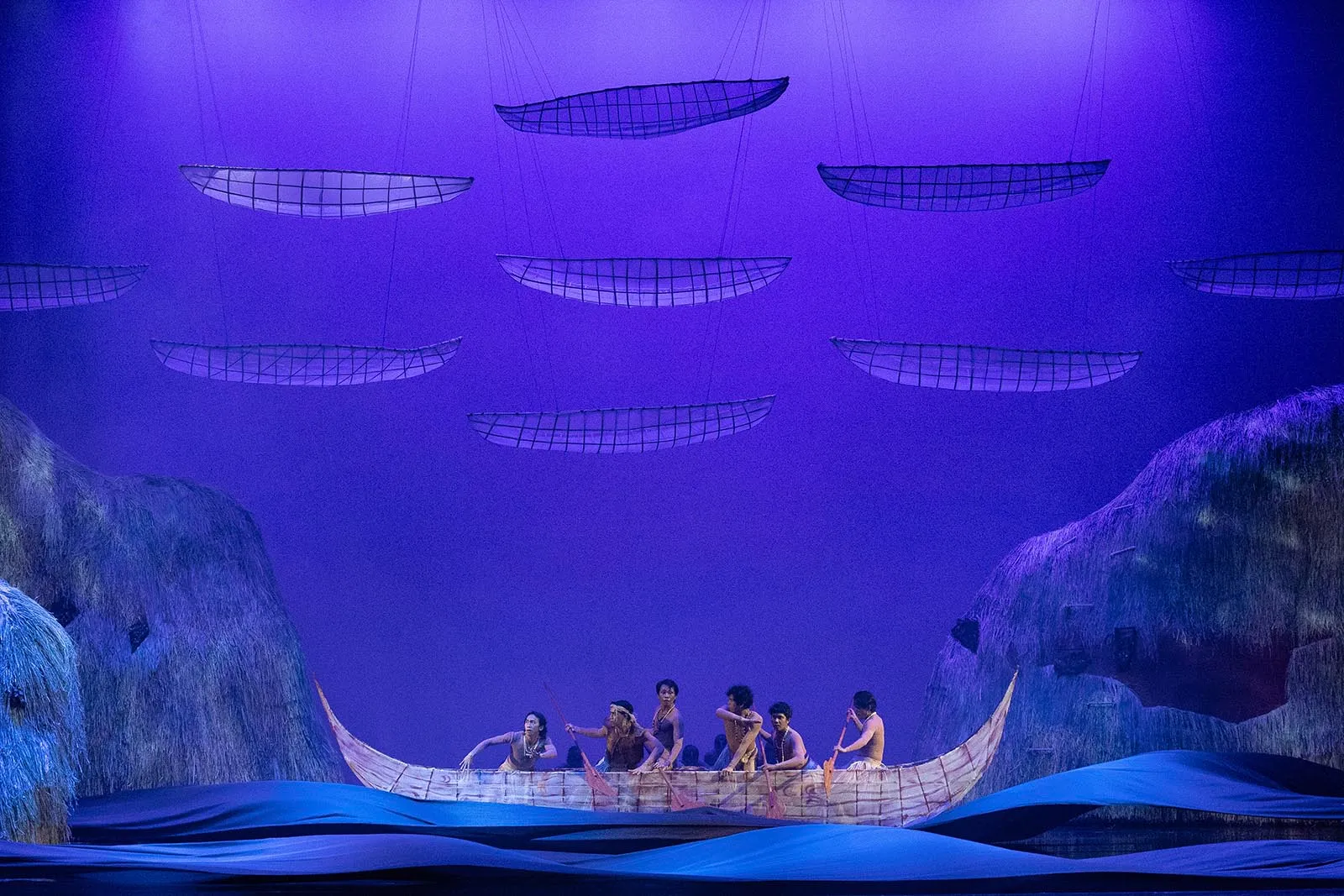 Stage scene with a long wooden canoe carrying dancers under blue lighting and suspended sail shapes above them.