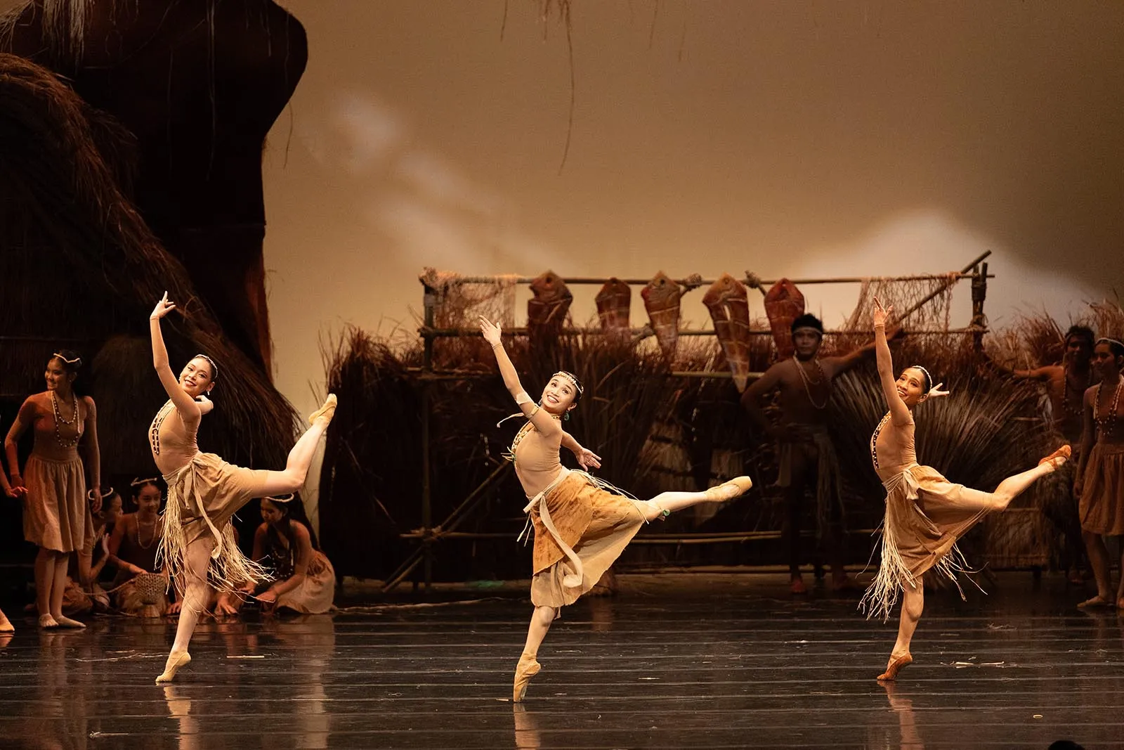 Three young ballerinas in tan fringe costumes perform a synchronized leap on a stage with a rustic tropical backdrop.