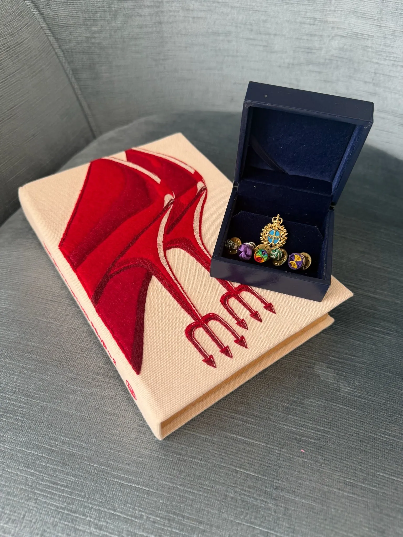 Open navy jewelry box containing a gold ring with a blue-turquoise centerpiece, placed next to a beige book with red stitched design on a gray couch.