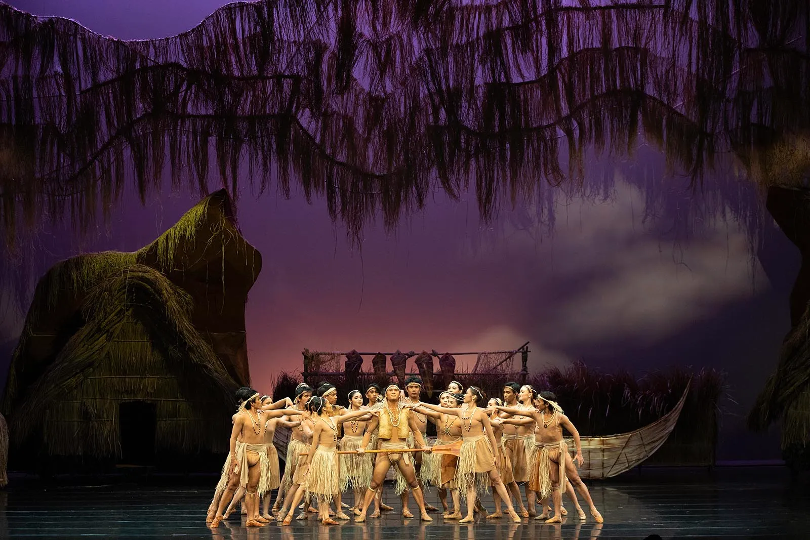 Group of dancers in grass skirts performing onstage with a canoe and tropical set behind them.