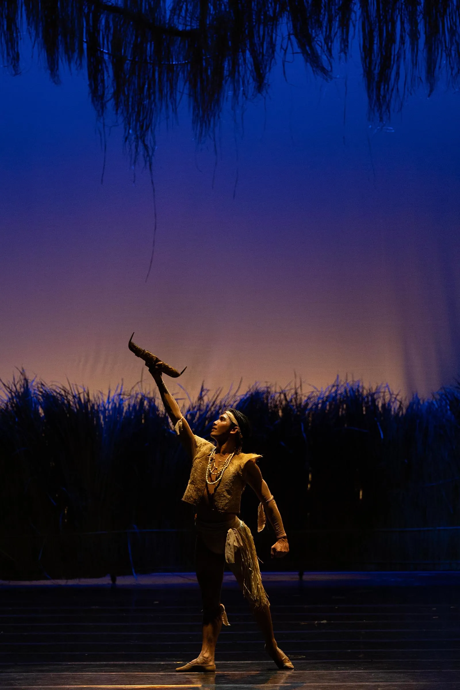 Dancer in fringed beige costume with beads raises a curved horn prop toward the sky on a dim blue-lit stage.