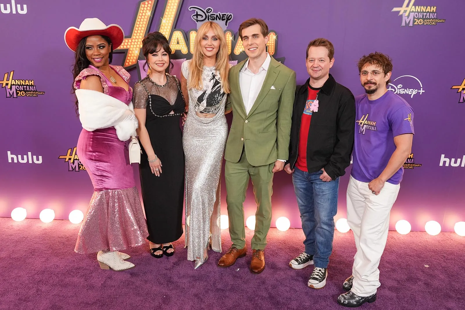 Six adults pose on a purple carpet at a Disney Hannah Montana event, wearing glamorous outfits with a branded backdrop behind them.