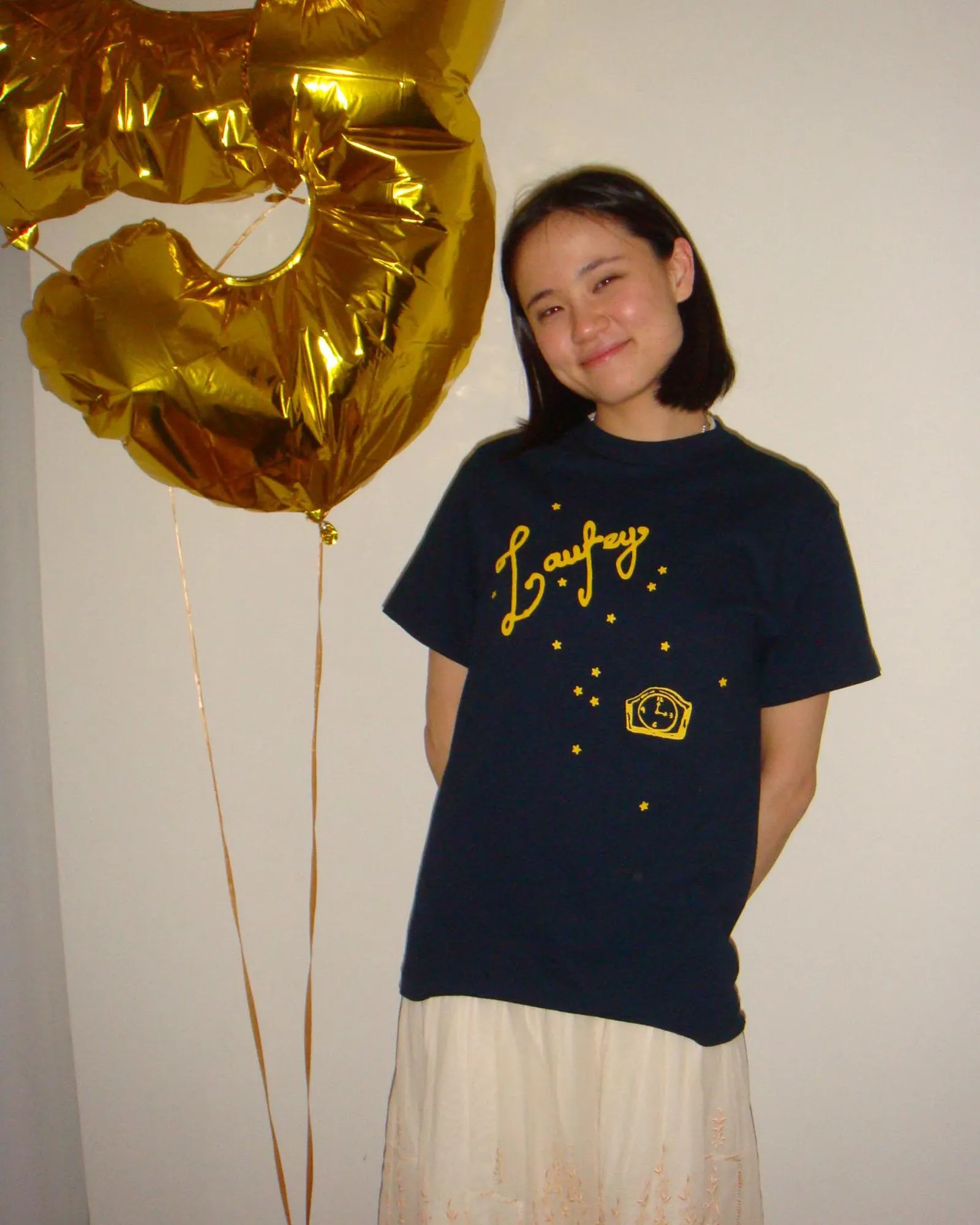 Smiling young woman in a navy T-shirt stands beside large gold foil balloons against a plain wall.