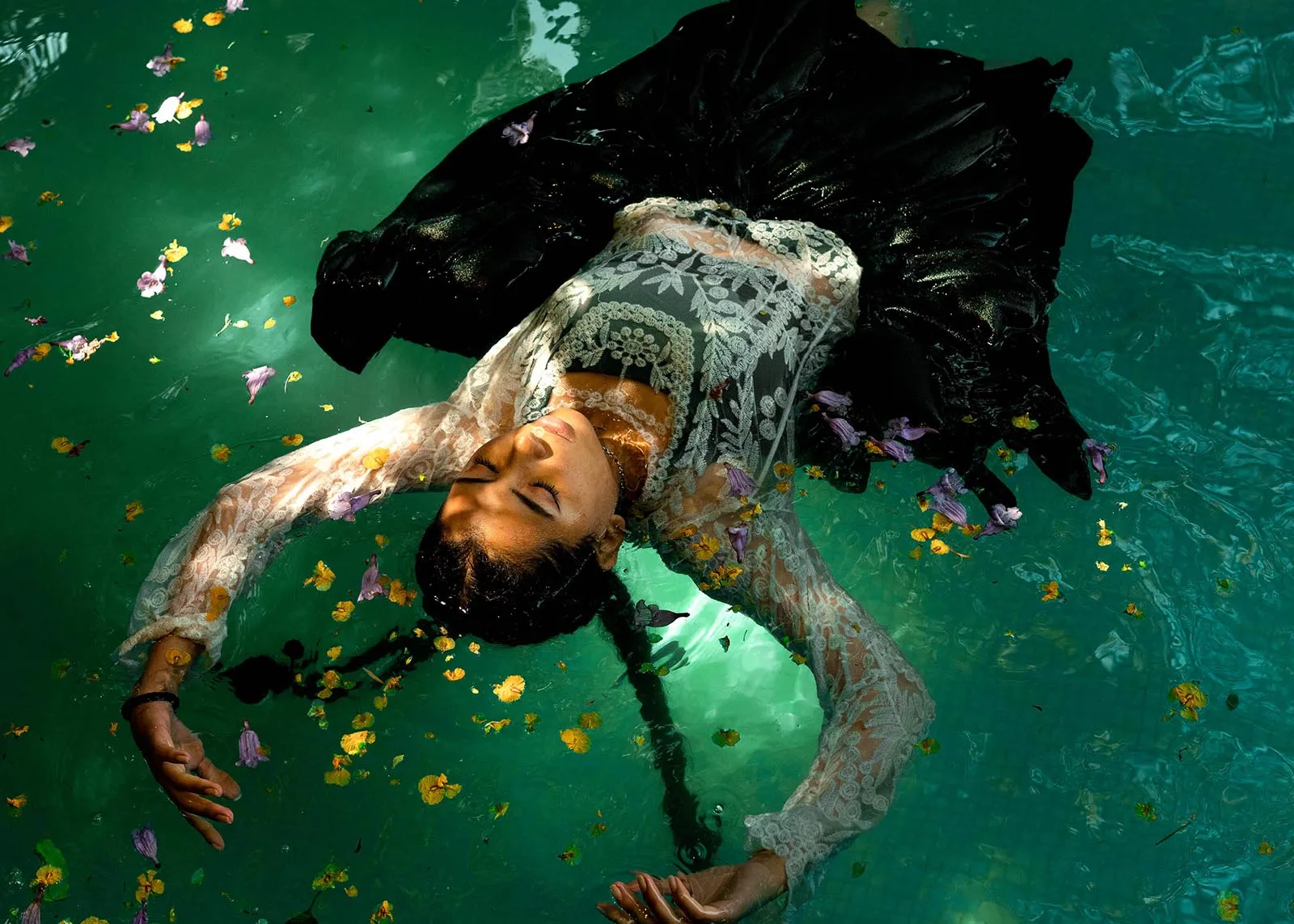Woman in a lace dress floats on her back in turquoise water, petals scattered around her.