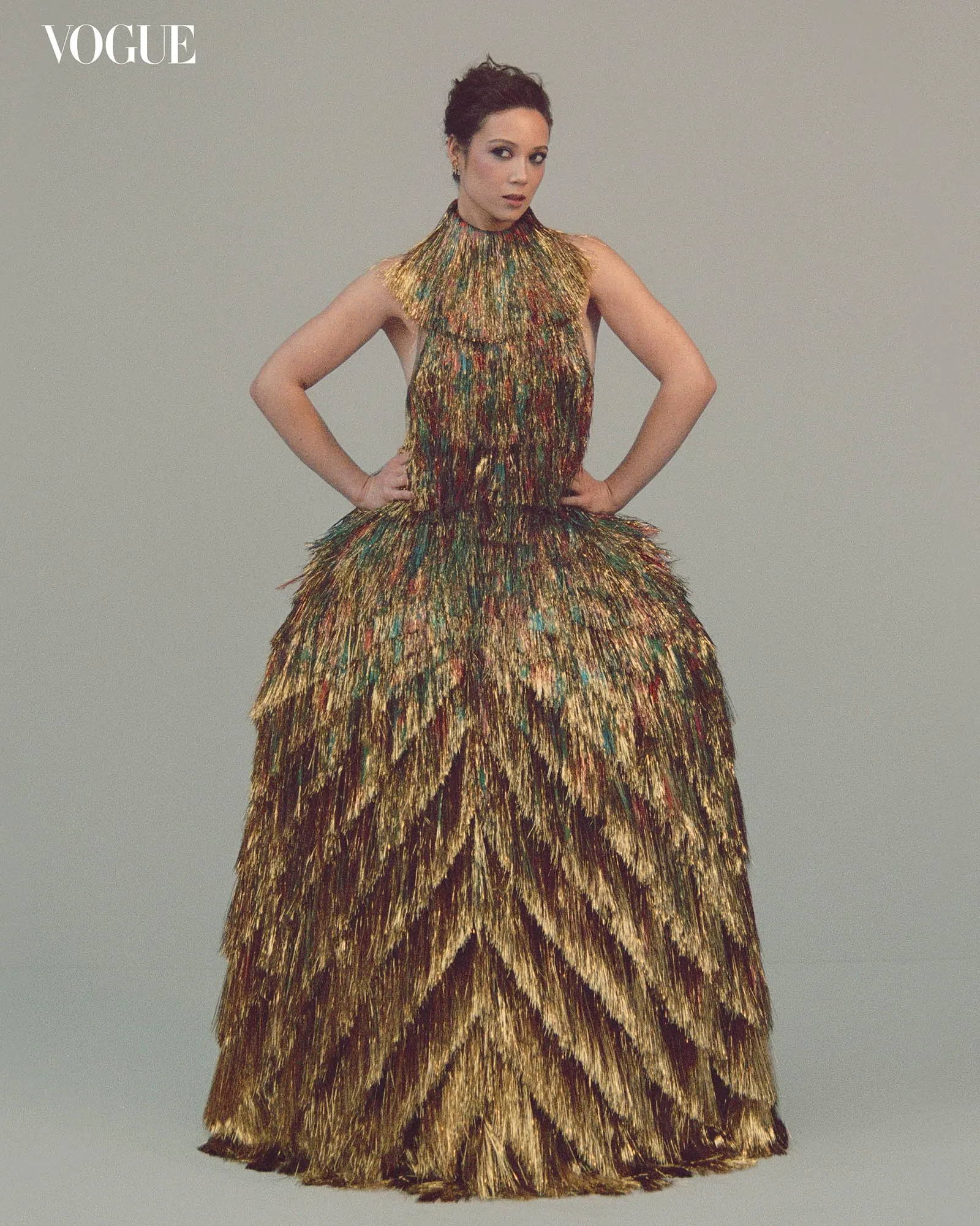 Model wearing a long multicolored feathered gown, standing with hands on hips; Vogue logo visible in top-left corner.