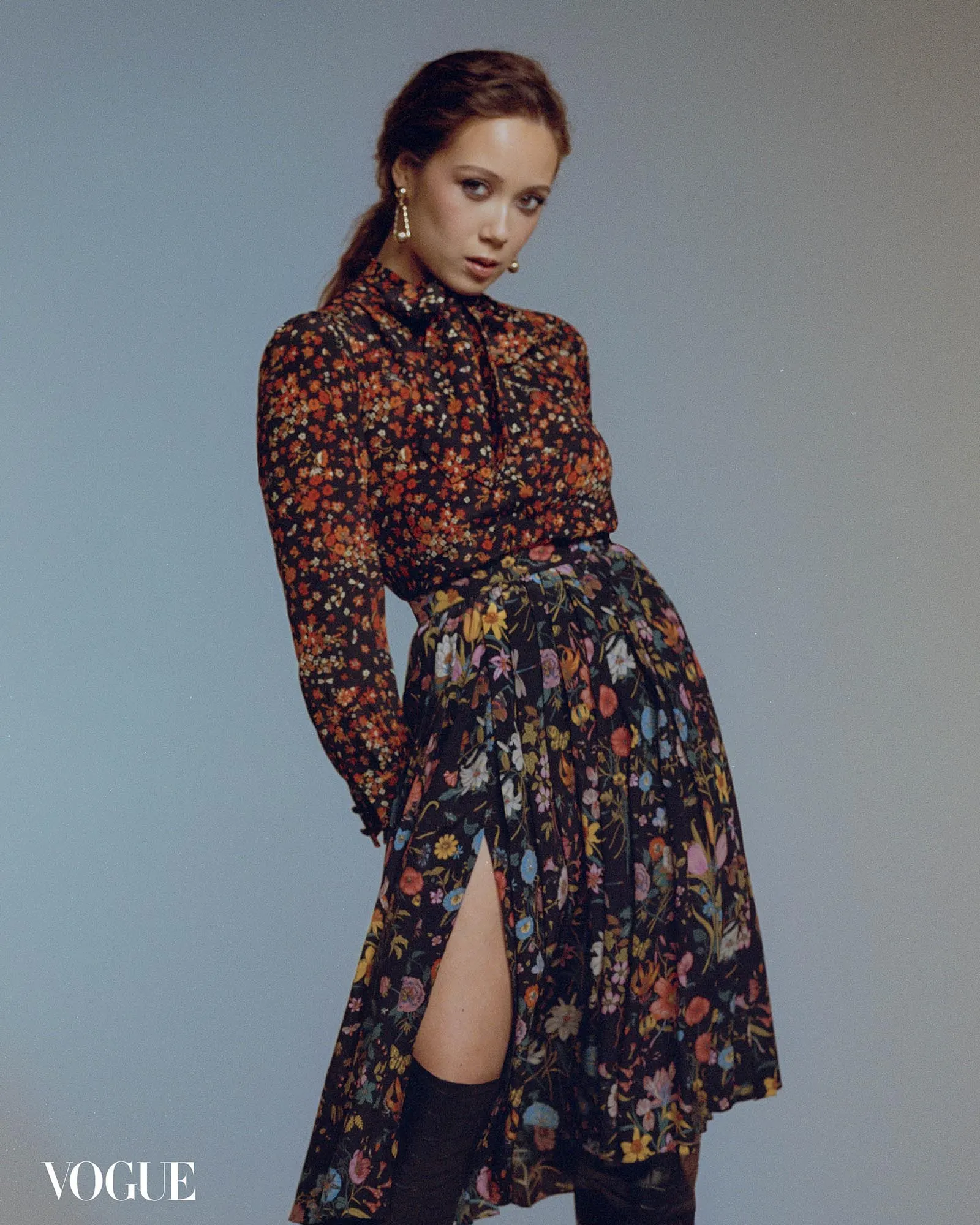 Model in a dark floral dress with long sleeves and a thigh-high slit, posing against a gray-blue gradient backdrop; Vogue logo visible in the corner.