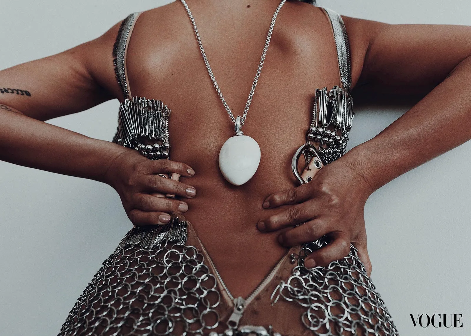 Back view of a model wearing a large white oval pendant necklace over a metallic, cut‑away bodice with chainmail details; Vogue logo in corner.