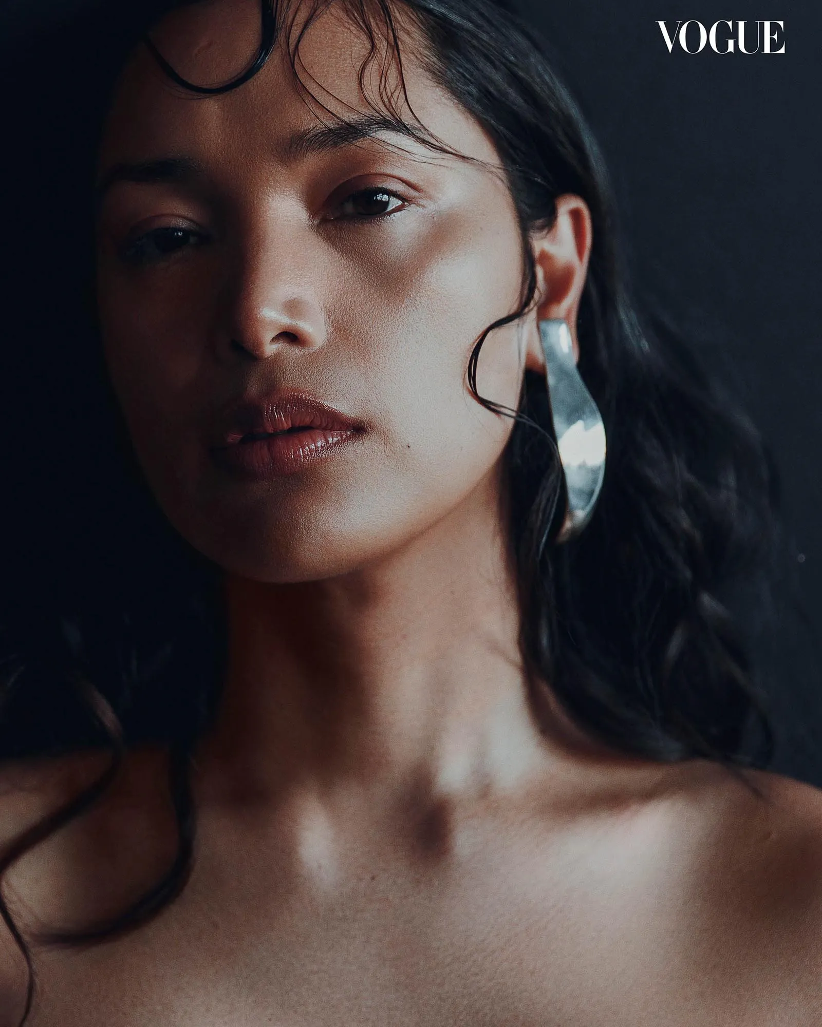 Close-up portrait of a woman with dark hair against a dark background; glossy lips and a silver earring visible; Vogue logo in the top-right corner.