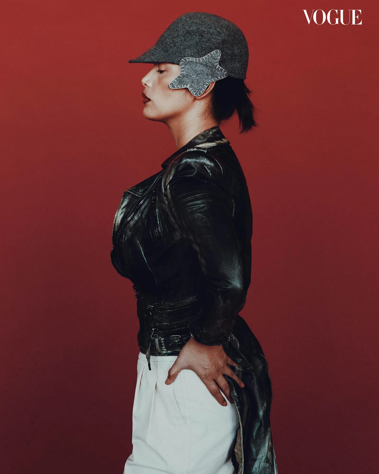 Side profile of a model wearing a gray hat with a star-shaped ear patch and a black leather jacket against a red background, Vogue styling.