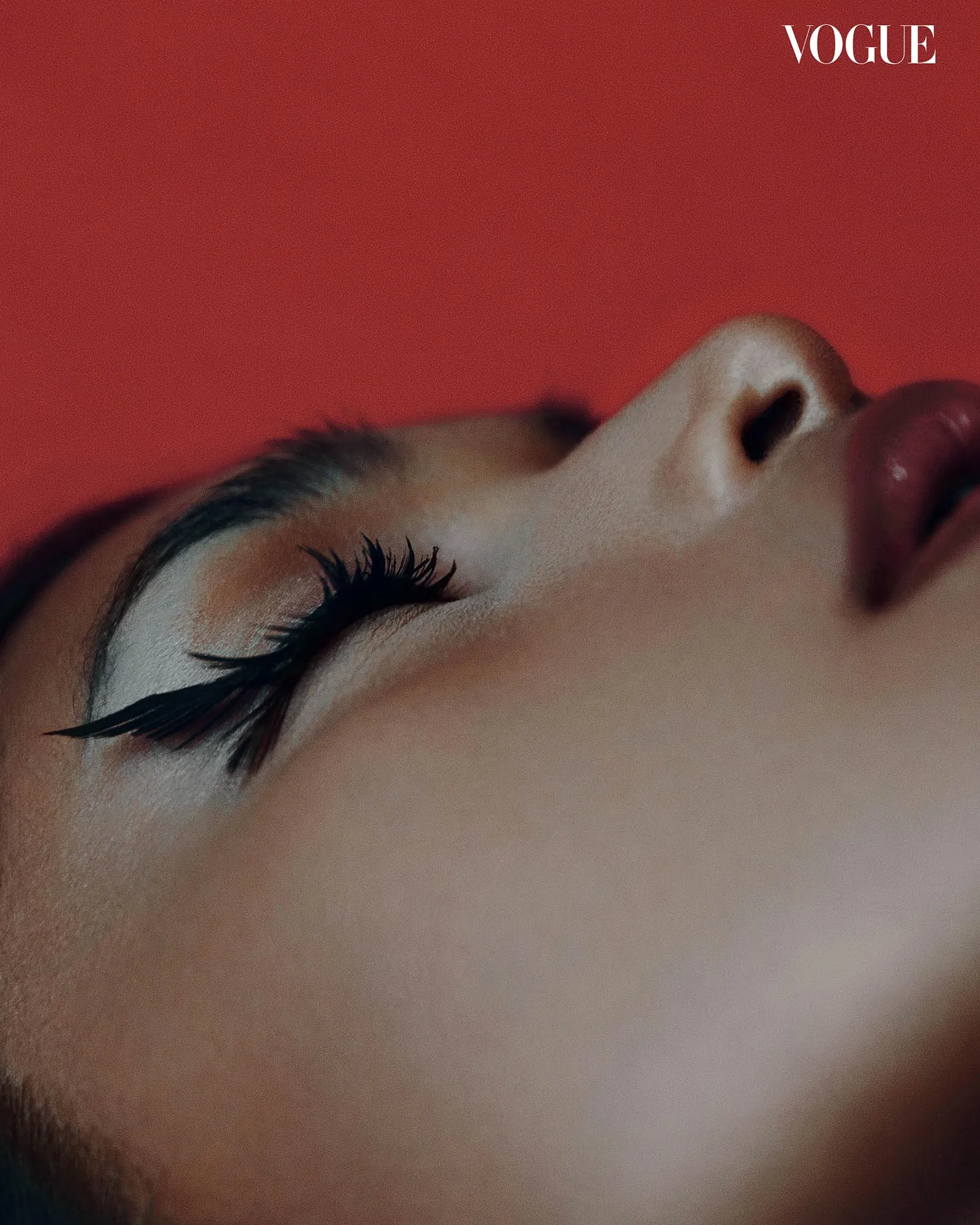 Close-up of a person's face tilted on a bold red background, with the Vogue logo in the top-right corner.