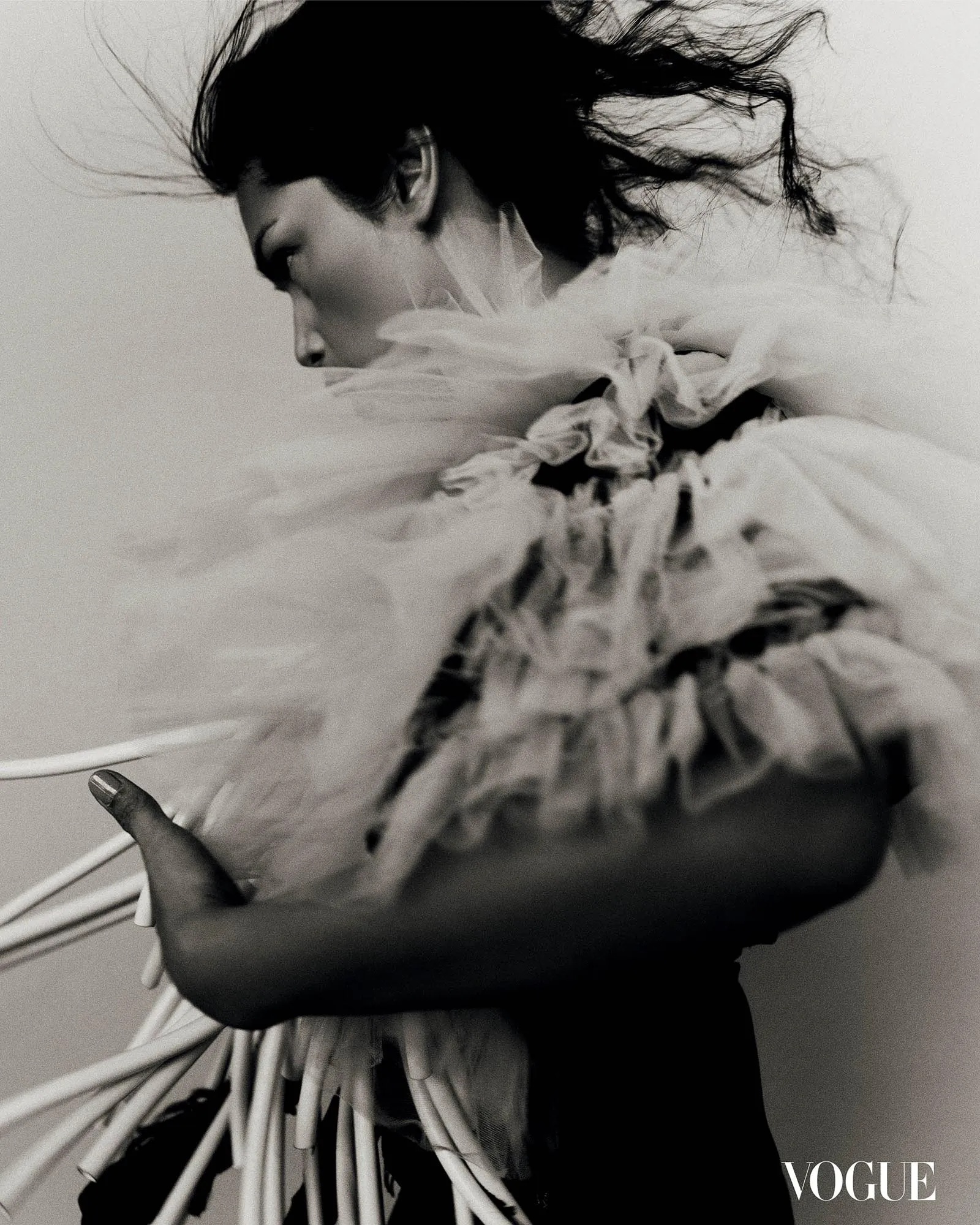 Editorial fashion shot: a woman in a dramatic, feathered garment in black and white, shown in profile with hair flowing; Vogue watermark visible.