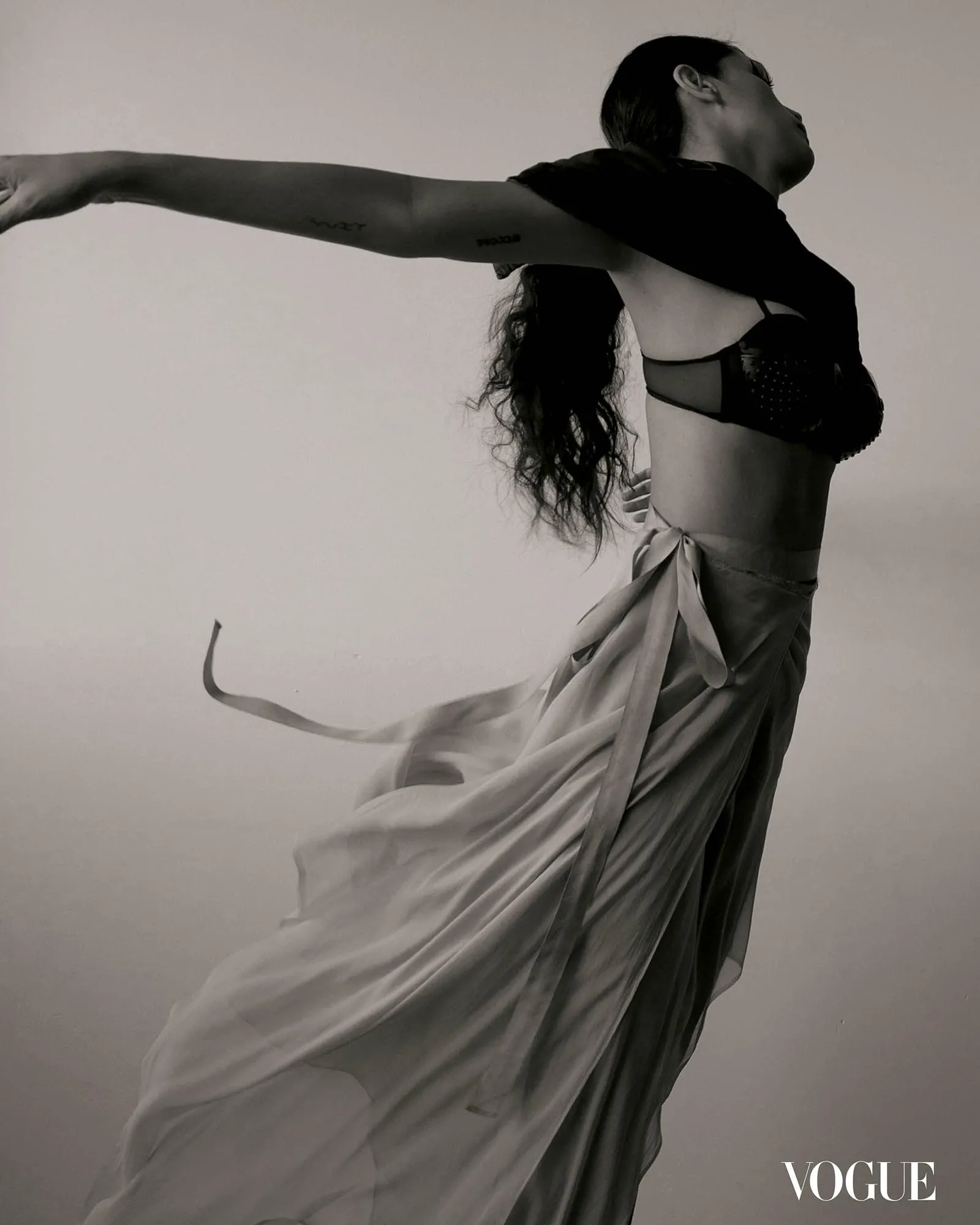 Black-and-white Vogue cover: model in a black bra with a flowing fabric, arms extended to the side, minimal background, Vogue logo bottom right.