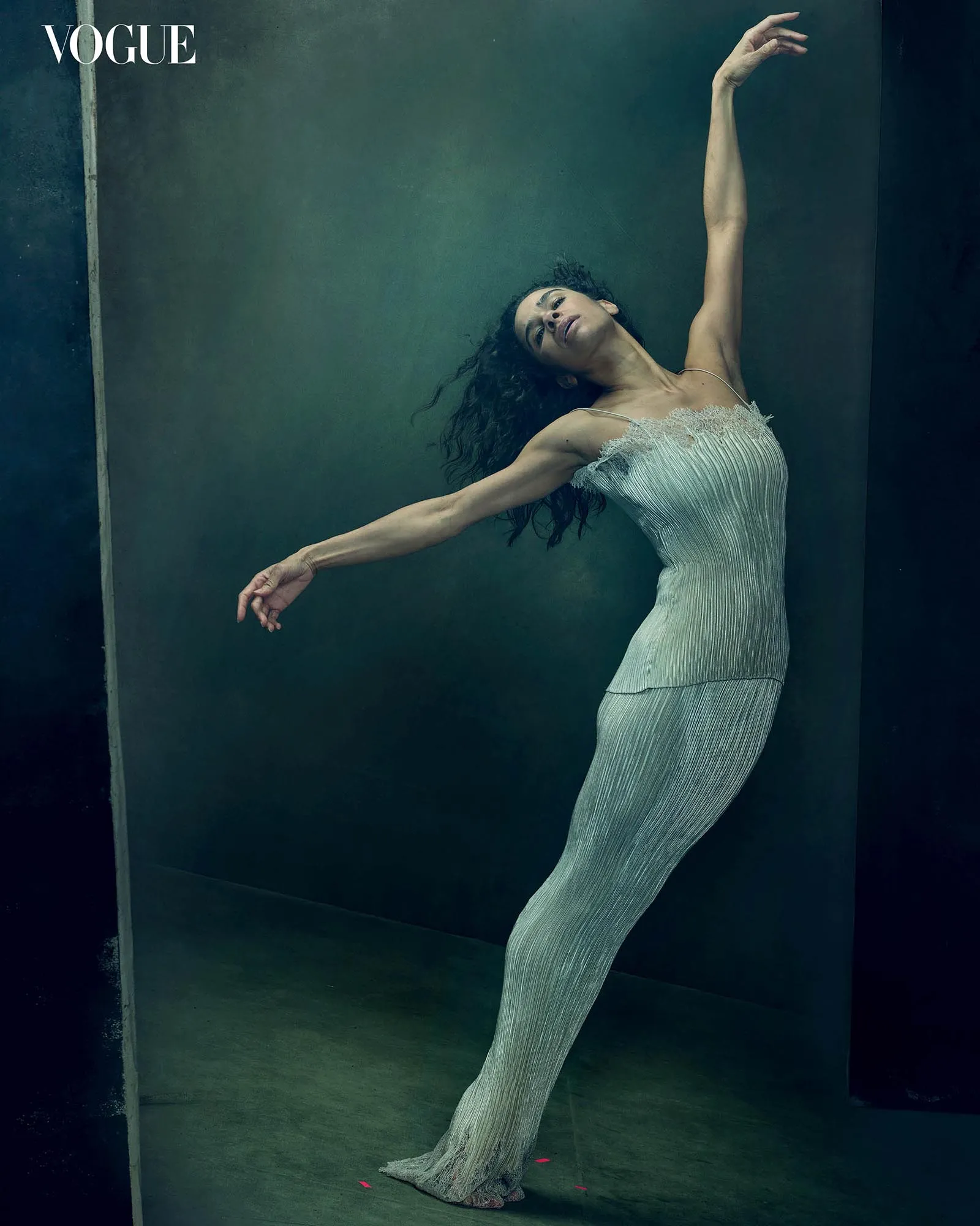 Model in a shimmering pleated gown leans diagonally with arms extended against a dark teal studio backdrop, Vogue logo in the top-left corner.