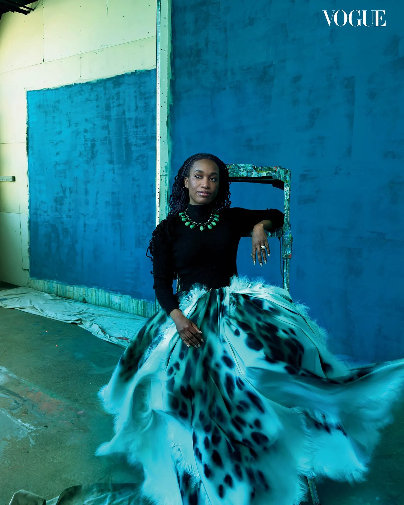 Fashion portrait: woman in a black top and blue patterned gown with a green beaded necklace, leaning on a blue-painted set.