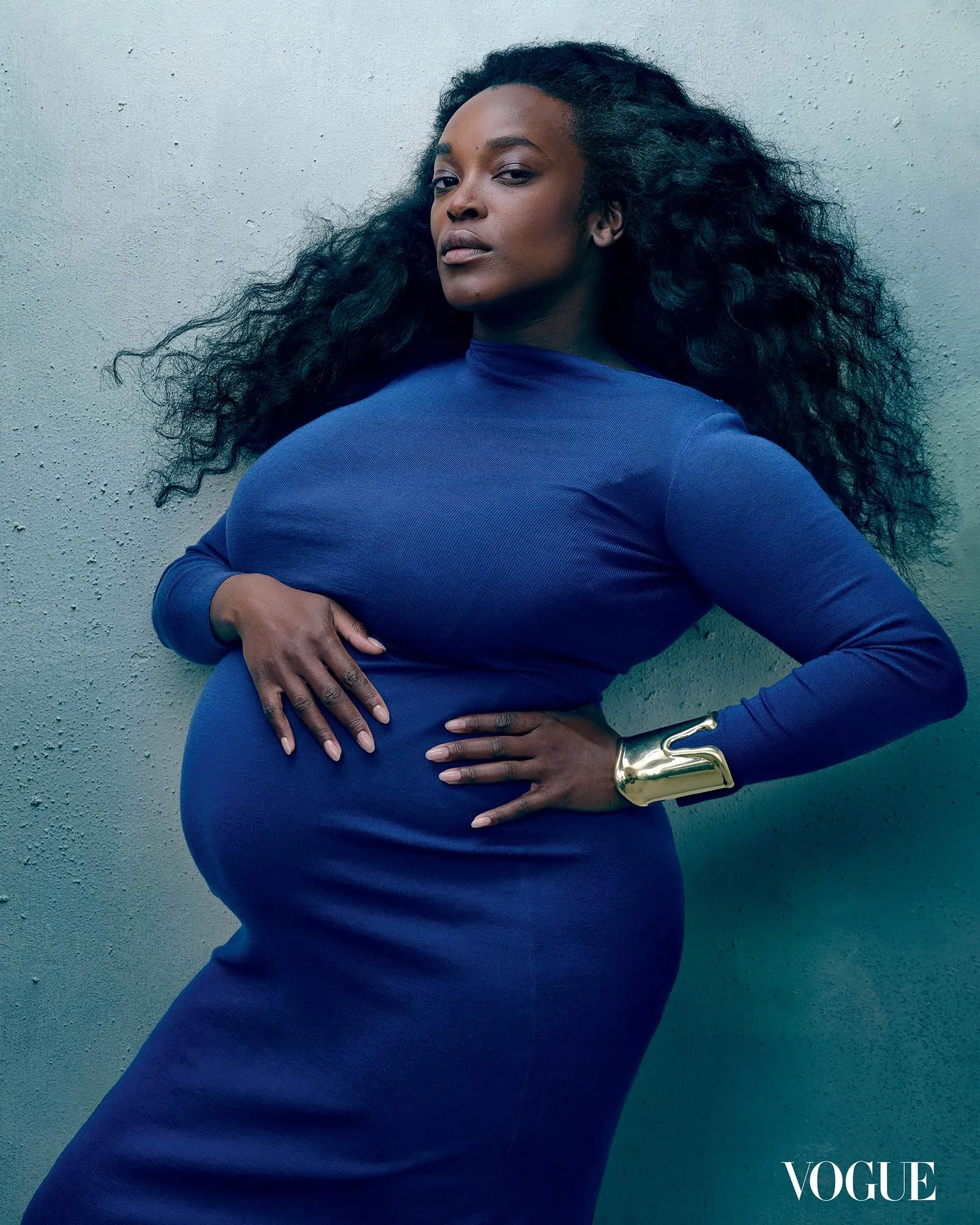 Pregnant woman in a blue dress leaning against a textured wall, Vogue watermark in the corner.
