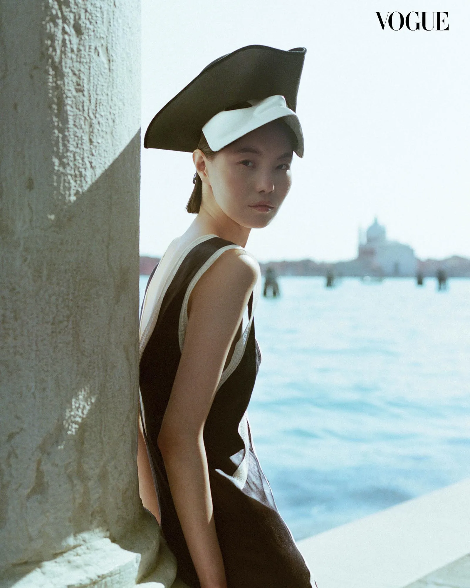 Woman in a black dress with a white bow hat leaning against a stone wall by the water, Vogue logo top-right.