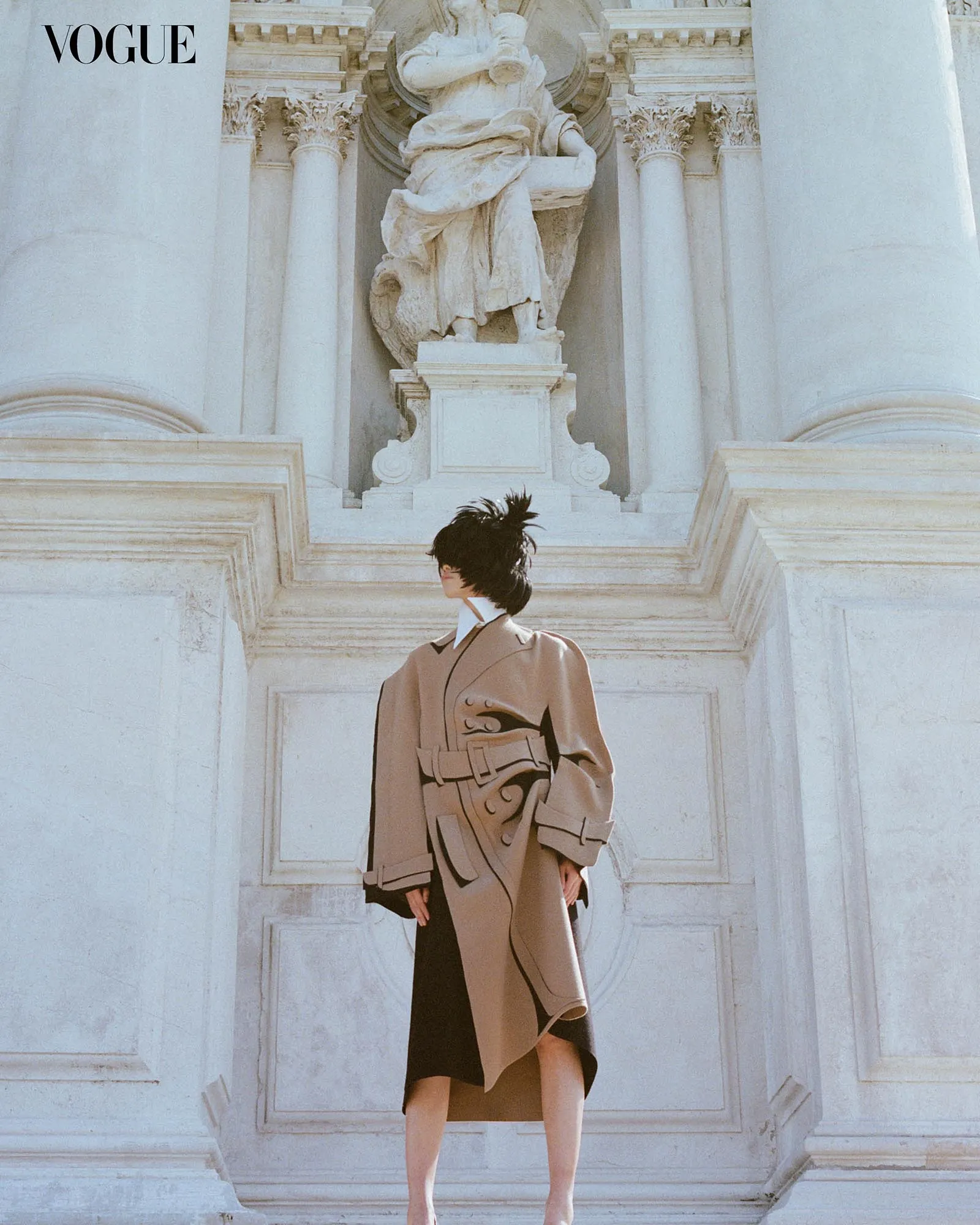 Vogue logo on a pale marble wall with a classical statue and columns; a model in a beige trench coat stands on the stone steps.