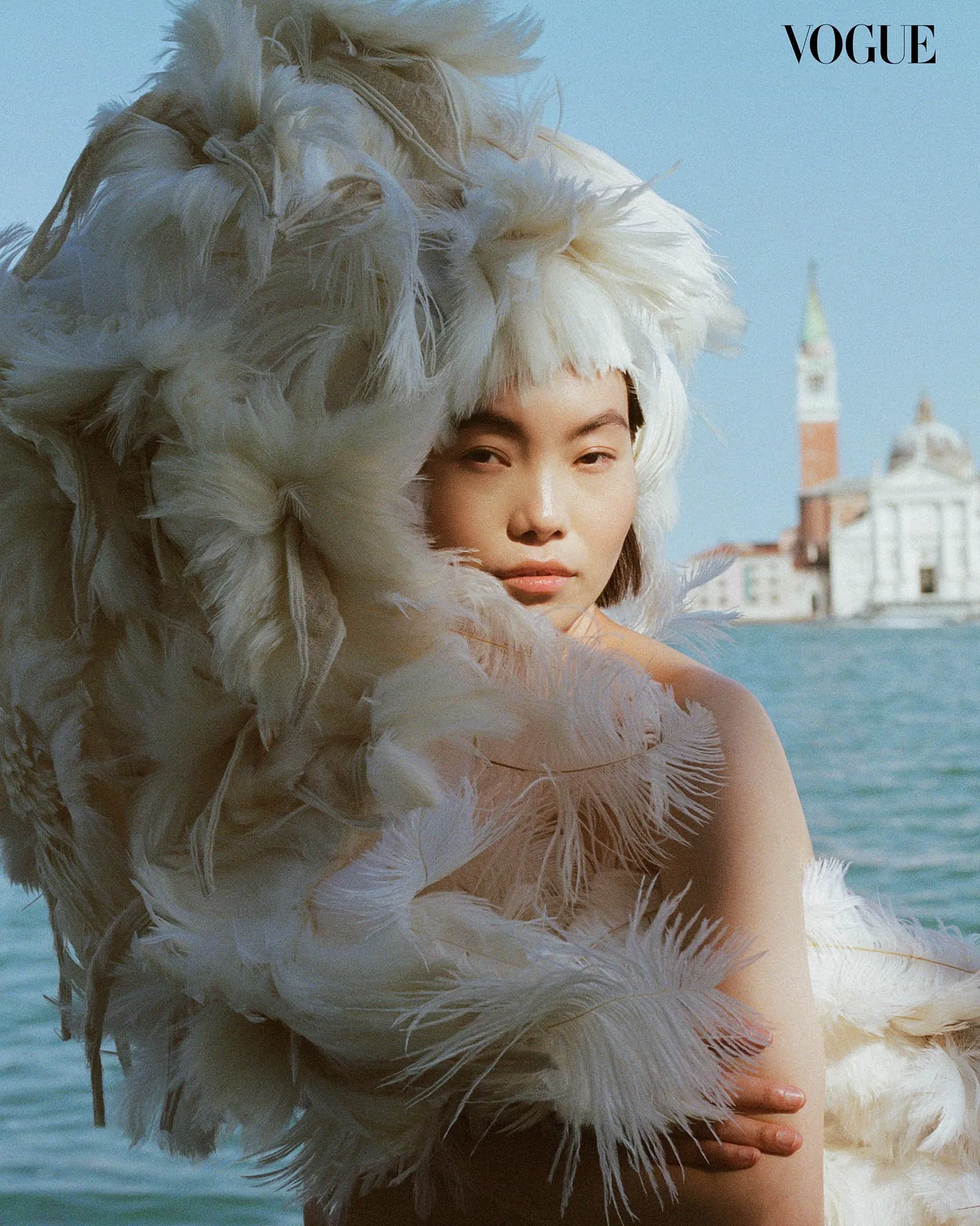 Model wrapped in large white feathers by the water, Vogue logo in the corner, bright blue sky behind.
