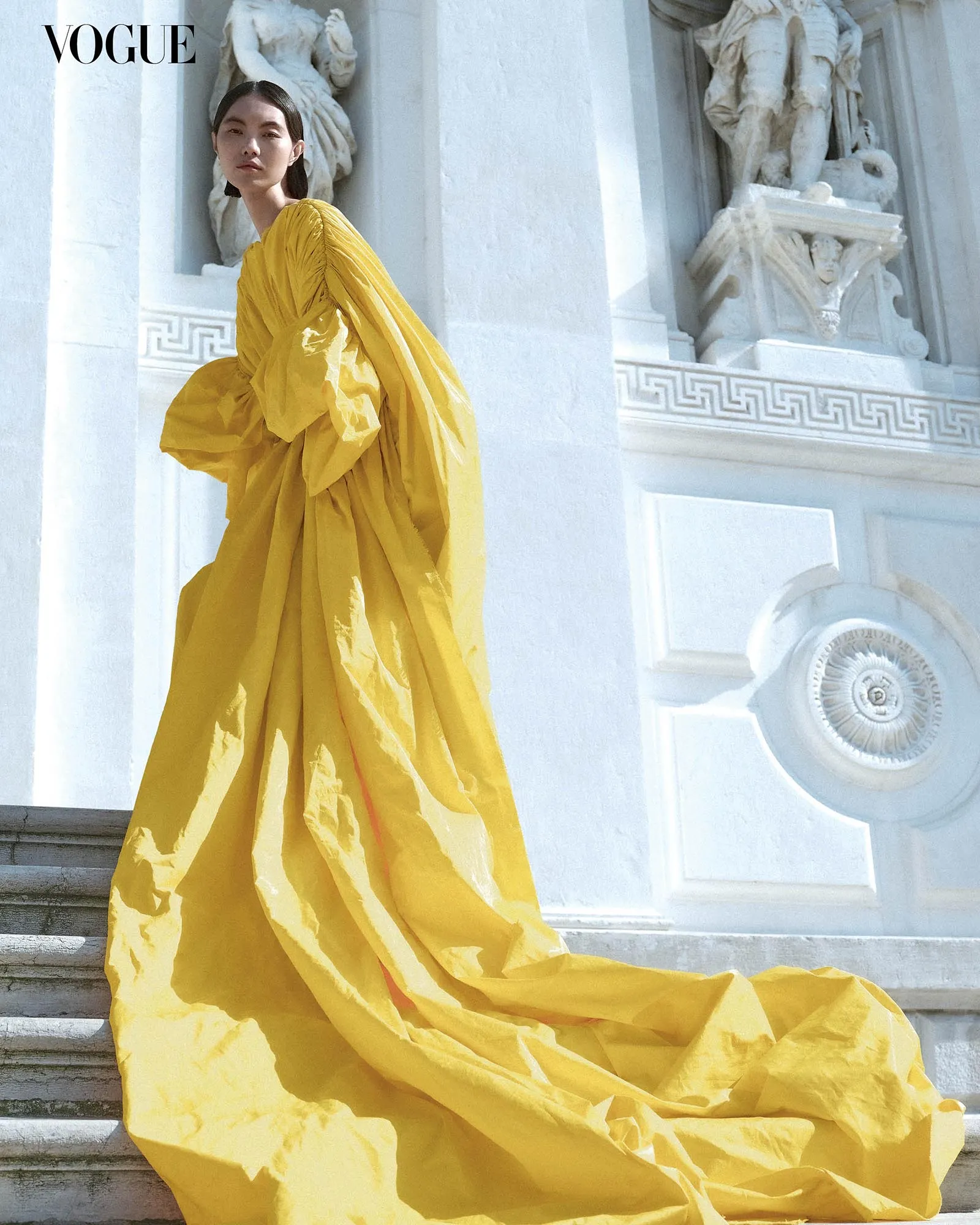 Model in a voluminous yellow gown stands on white steps beside ornate architectural statues; Vogue logo top left.