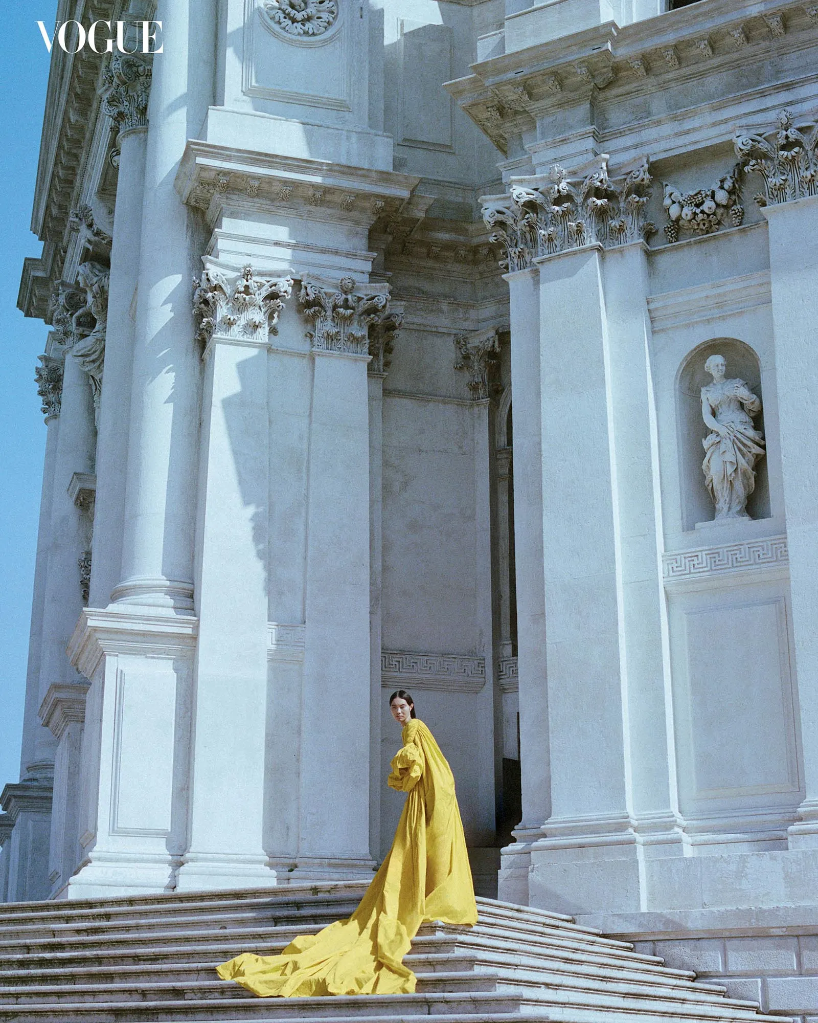 Woman in a flowing yellow dress standing on marble steps beside a white neoclassical building with ornate pillars; Vogue logo appears in top-left.