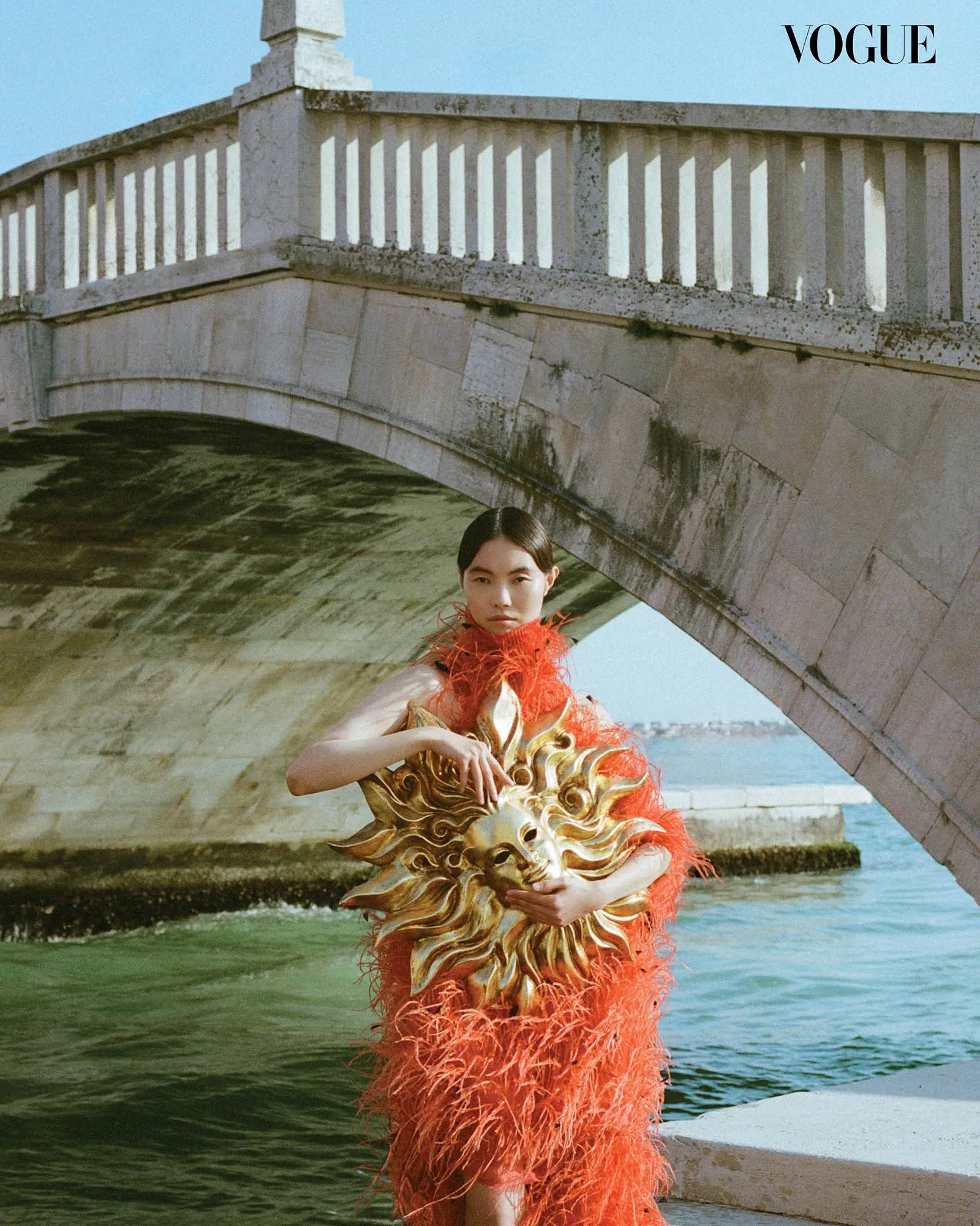 Fashion model in an orange feathered dress stands under a stone bridge by the water; Vogue logo in the corner.